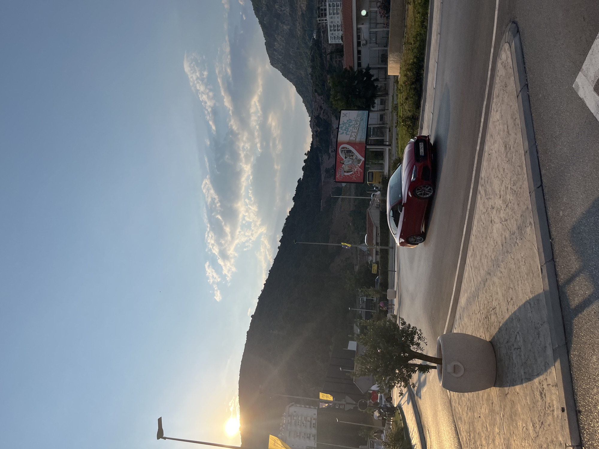 A red car is parked on the side of a road next to a sidewalk with a large planter containing a tree. The scene is bathed in the warm glow of the setting sun, which is visible low on the horizon, casting long shadows. A steep, tree-covered mountain dominates the background, with buildings nestled at its base and along the roadside. The sky is a clear blue with wispy clouds. A billboard is visible near the mountain. The overall mood is serene and peaceful, indicative of a late afternoon or early evening in a coastal town. The location context suggests Budva, Montenegro.