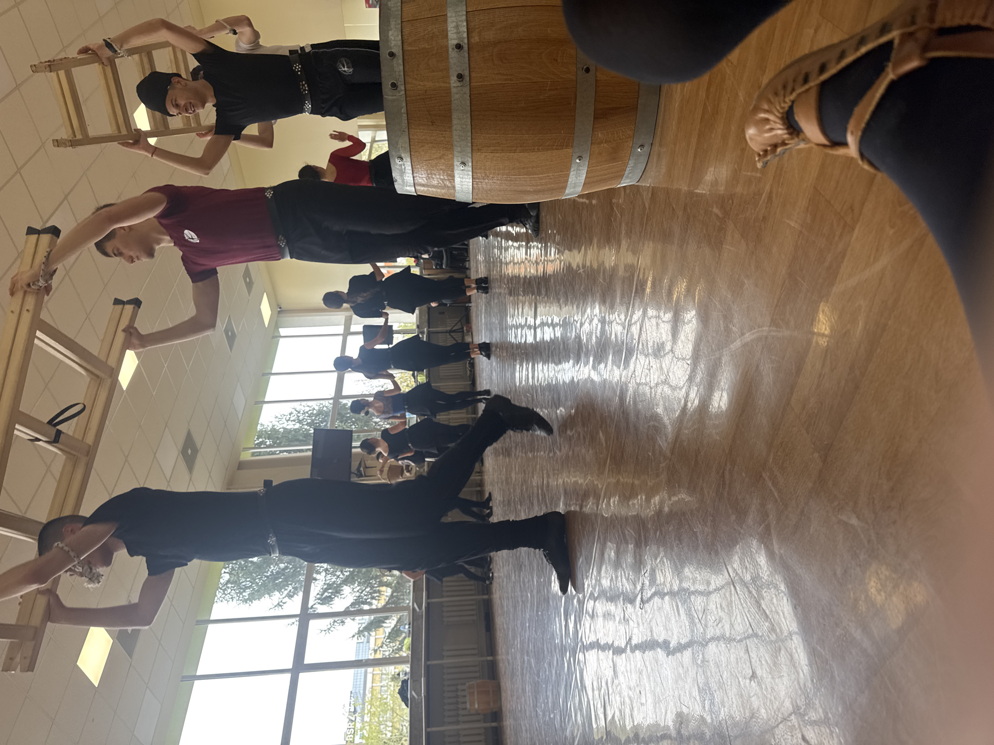 The image captures an indoor scene, possibly a dance or exercise studio, with a group of individuals engaged in what appears to be a choreographed routine involving ladders. The studio has large windows providing natural light and a glossy wooden floor. In the foreground, a person wearing black leggings and brown ballet flats is partially visible. The people in the scene are dressed in athletic wear, performing actions related to the ladders and a large wooden barrel. The photograph has been taken at an angle that causes distortion. The location cannot be determined to be Sofia, Bulgaria based only on this image.