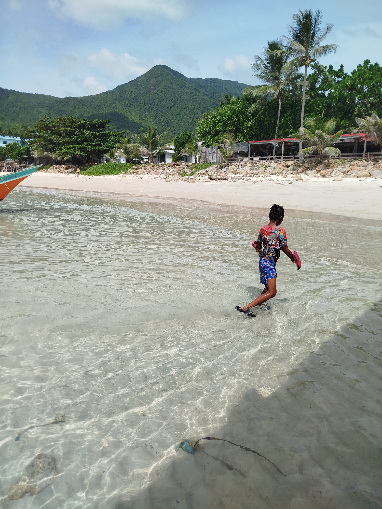 A young person walks in shallow, clear water near a sandy beach on Ko Pha-ngan District, Thailand. The person is wearing a colorful t-shirt and blue shorts, with pink flip-flops in their hands and dark footwear on their feet. The water is slightly rippled, revealing the sandy bottom. In the background, a white sand beach is lined with rocks, palm trees, and small buildings with red roofs. Lush green hills rise in the distance under a partly cloudy blue sky. A portion of an orange and turquoise boat is visible on the left side of the frame.