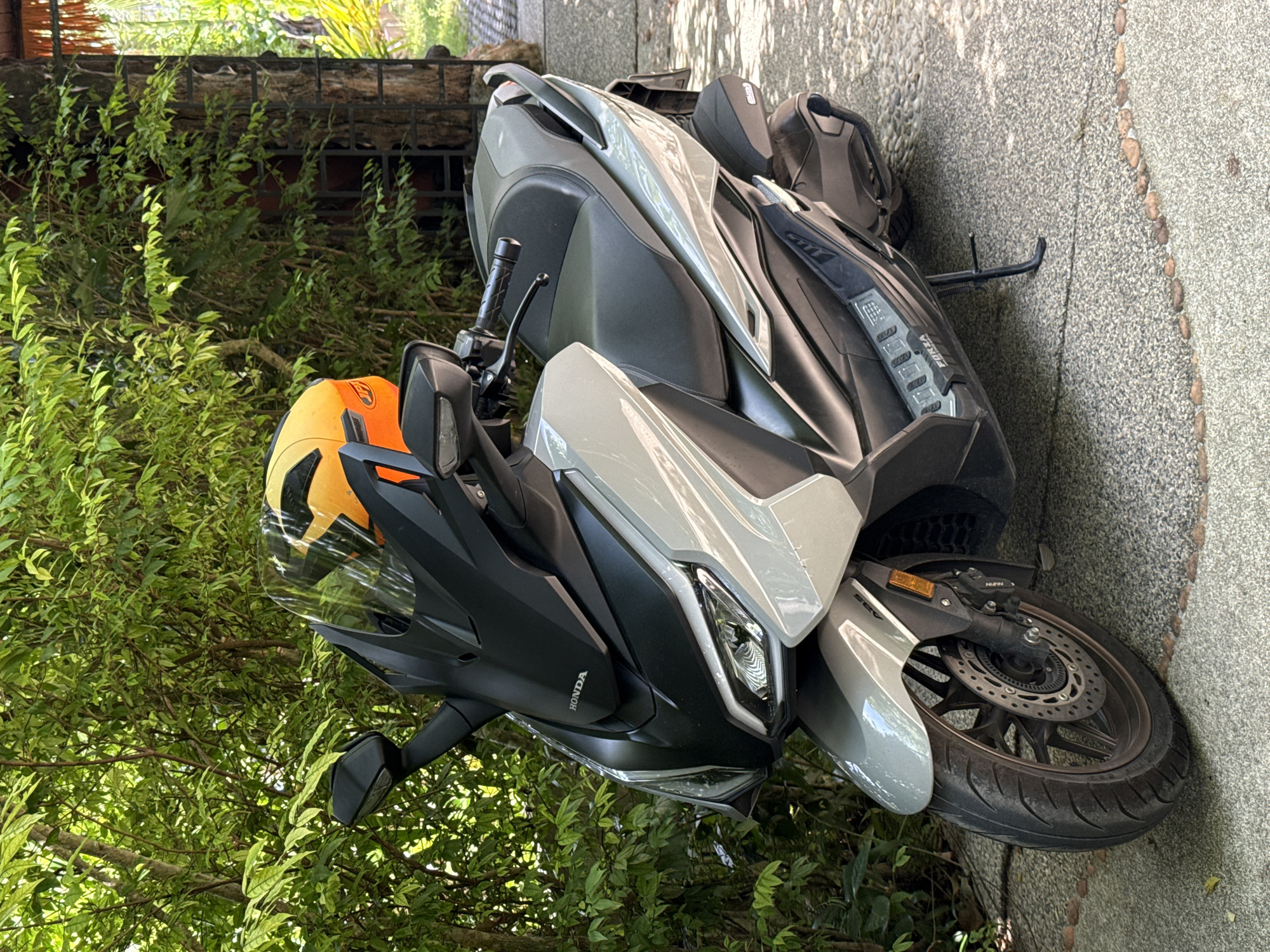 A light gray Honda Forza scooter is parked on a pebble-strewn pathway in a tropical setting, likely a resort or private residence, in Ko Samui District, Thailand. The scooter is facing the viewer at a slight angle, with its front wheel on the left. An orange and black helmet rests on the scooter's windshield. Behind the scooter, lush green foliage provides a dense backdrop, with sunlight filtering through the leaves. To the right of the scooter, part of a wooden structure with thatched roofing is visible, suggesting an outdoor covered area. The ground is a textured surface composed of small pebbles and some larger, rounded stones forming a border. The scooter's features, including the "HONDA" logo on the front, the "FORZA" branding on the footboard, and the ABS marking on the front fender, are discernible. No people are present in the scene.