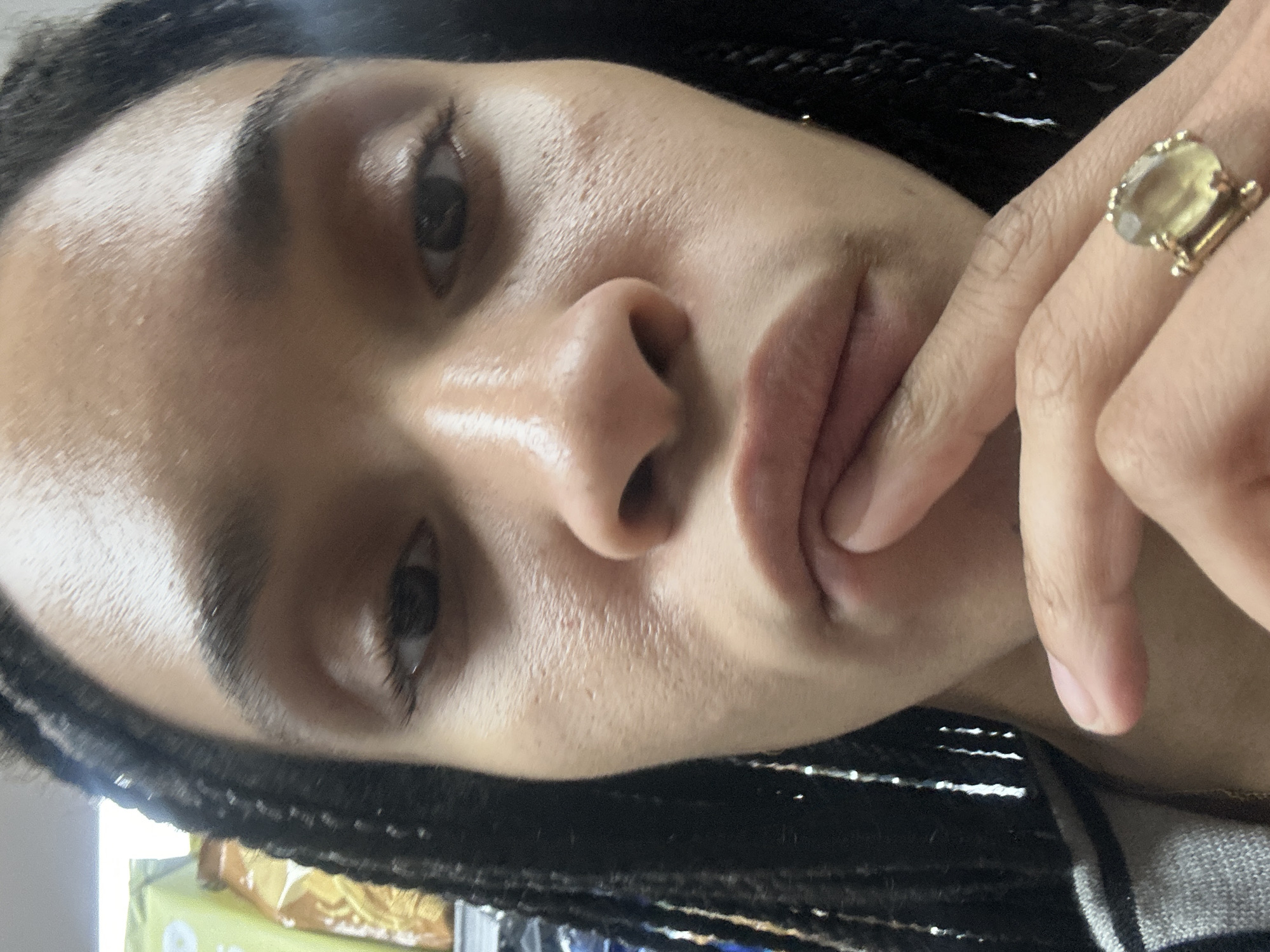 A close-up, slightly angled portrait shows a person with dark eyes and braided hair. The individual's skin has a visible texture. The person's hand is raised, with a finger touching their lower lip. On their finger, there is a gold ring with a yellow stone. In the blurry background, there are objects including what appears to be a package and a striped garment. There is no location information to confirm this is in the city of London, United Kingdom.