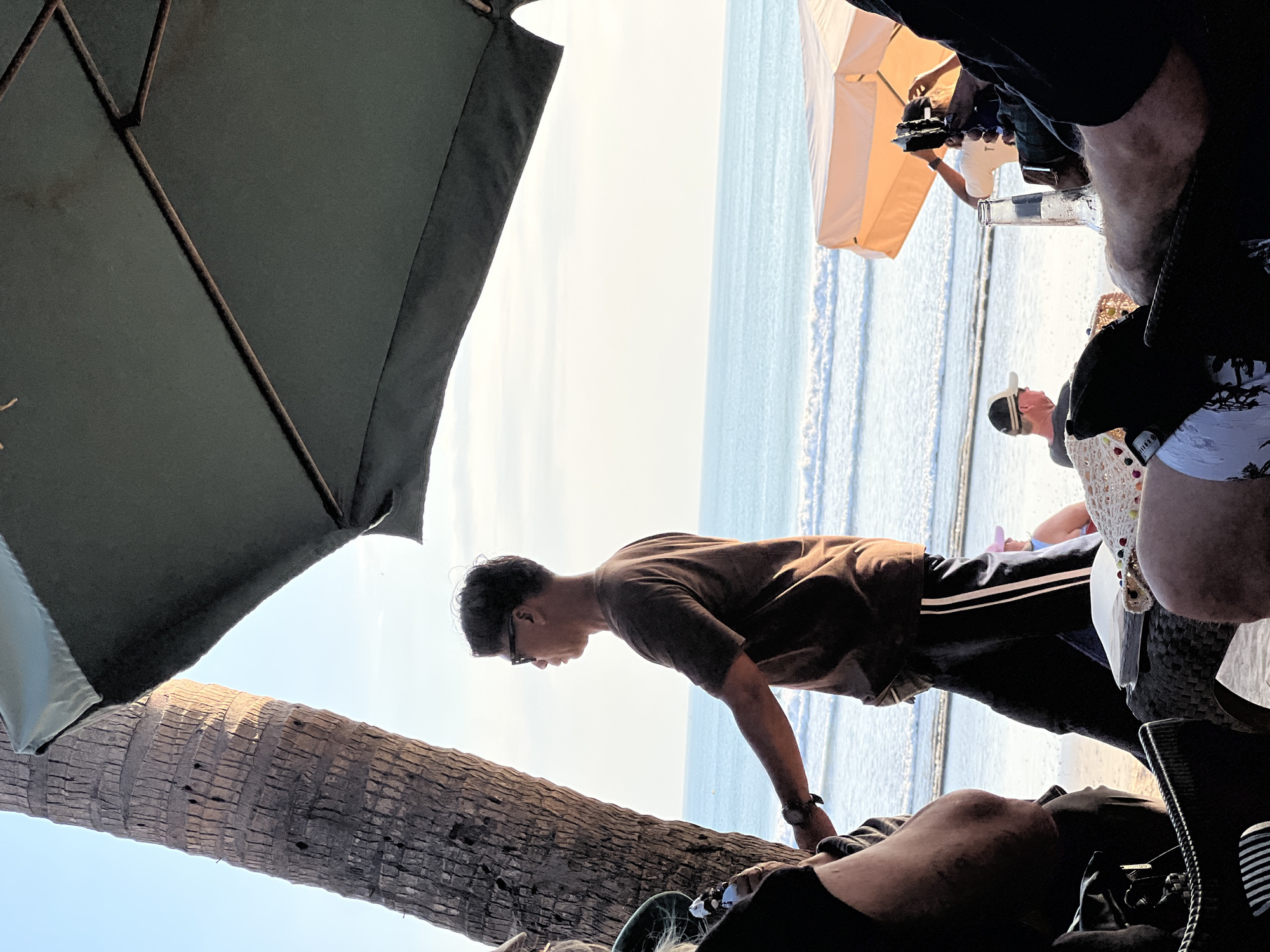 In Kuta, Indonesia, a scene unfolds under the shade of a large green umbrella. A man with dark hair and sunglasses, wearing a brown t-shirt and black pants with white stripes, stands near a palm tree, looking out at the ocean. The sea is calm with gentle waves, and the sky is clear. In the background, beachgoers relax under a light-colored umbrella, and a bottle of beer sits on a table. The image captures a peaceful moment at the beach, with the sounds of the waves and the warmth of the sun.