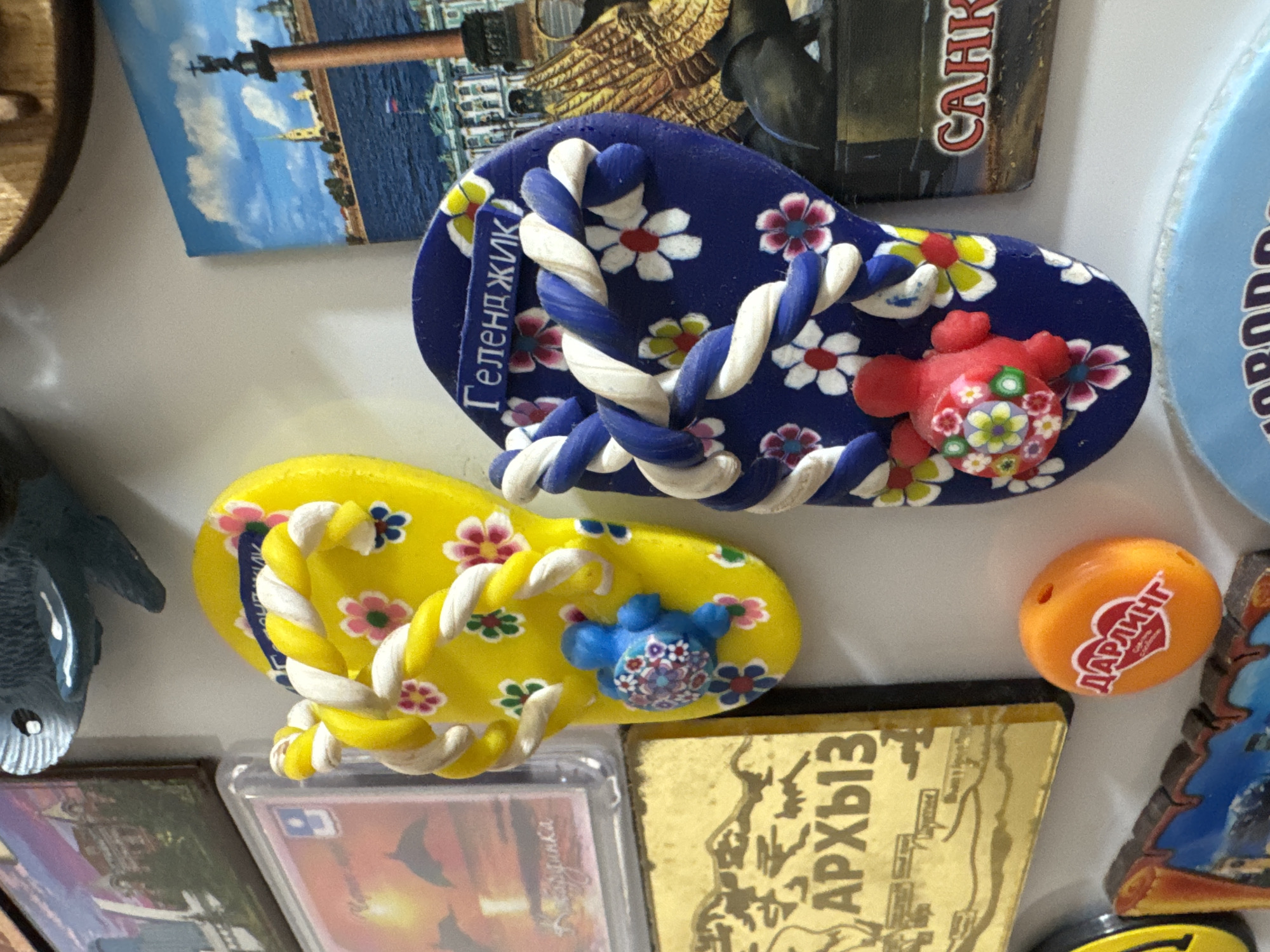 The image shows a collection of refrigerator magnets. In the center are two flip-flop magnets decorated with flowers and turtles. One is yellow with blue and white straps, and the other is blue with white straps. To the left of the flip-flops is a yellow magnet with a map-like design and text in Cyrillic. To the right of the blue flip-flop is a magnet with a picture of a building and water, and text that appears to say "САНК". Below the yellow flip-flop is an orange, oval-shaped magnet with a heart symbol and Cyrillic text. The overall scene is a close-up of a refrigerator door covered in magnets. There are no people visible in the image. The location of Mikhaylovsk, Russia is not ascertainable from the visual information presented in the image.