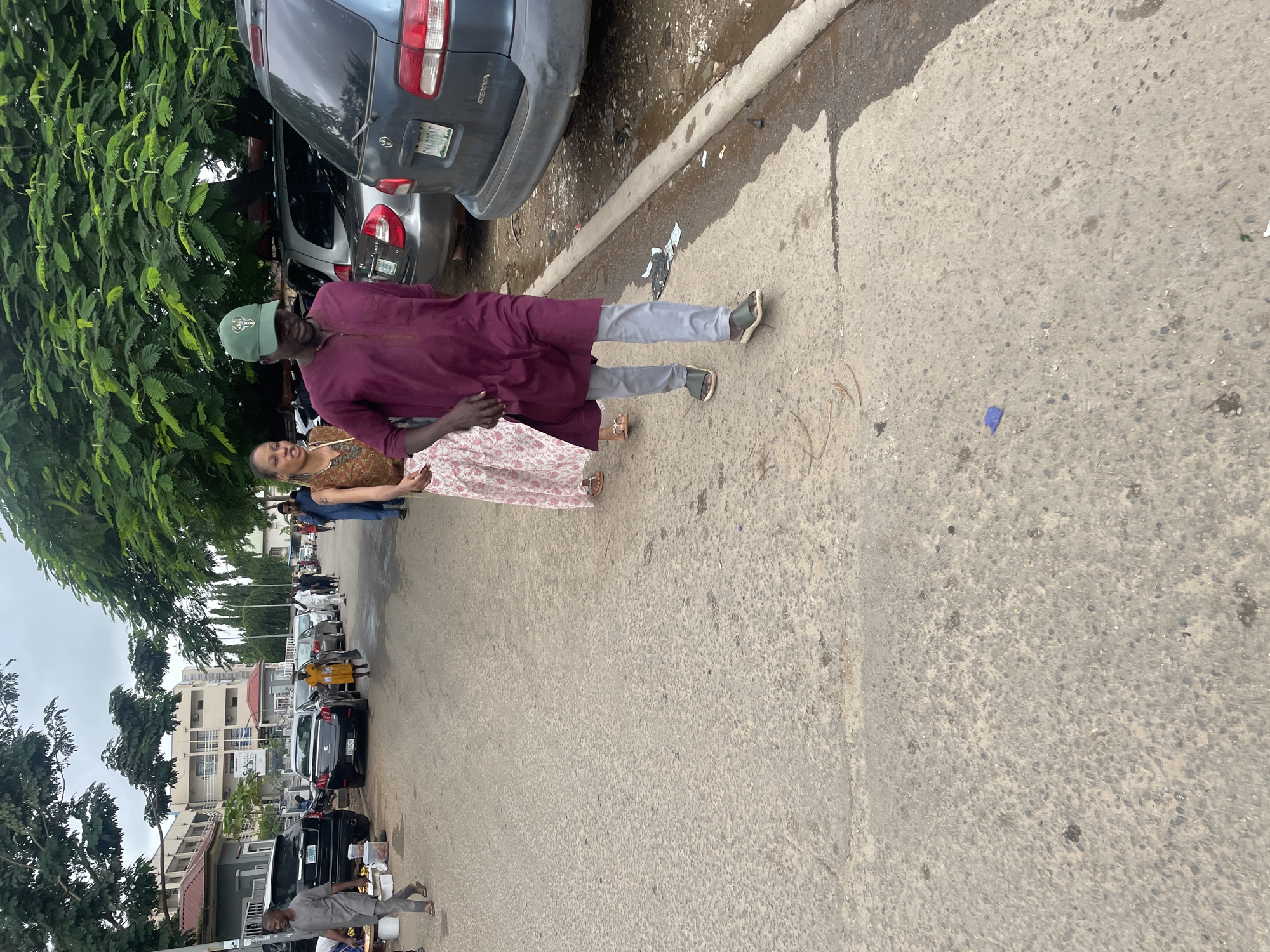 A man in a burgundy tunic and light gray pants walks on a paved road in Abuja, Nigeria, wearing a green cap with a deer logo. He is accompanied by a woman in a floral print skirt and a brown top. Several vehicles are parked along the road, including a dark blue minivan, a silver car, and a black SUV. Buildings and trees line the street. The weather appears overcast.