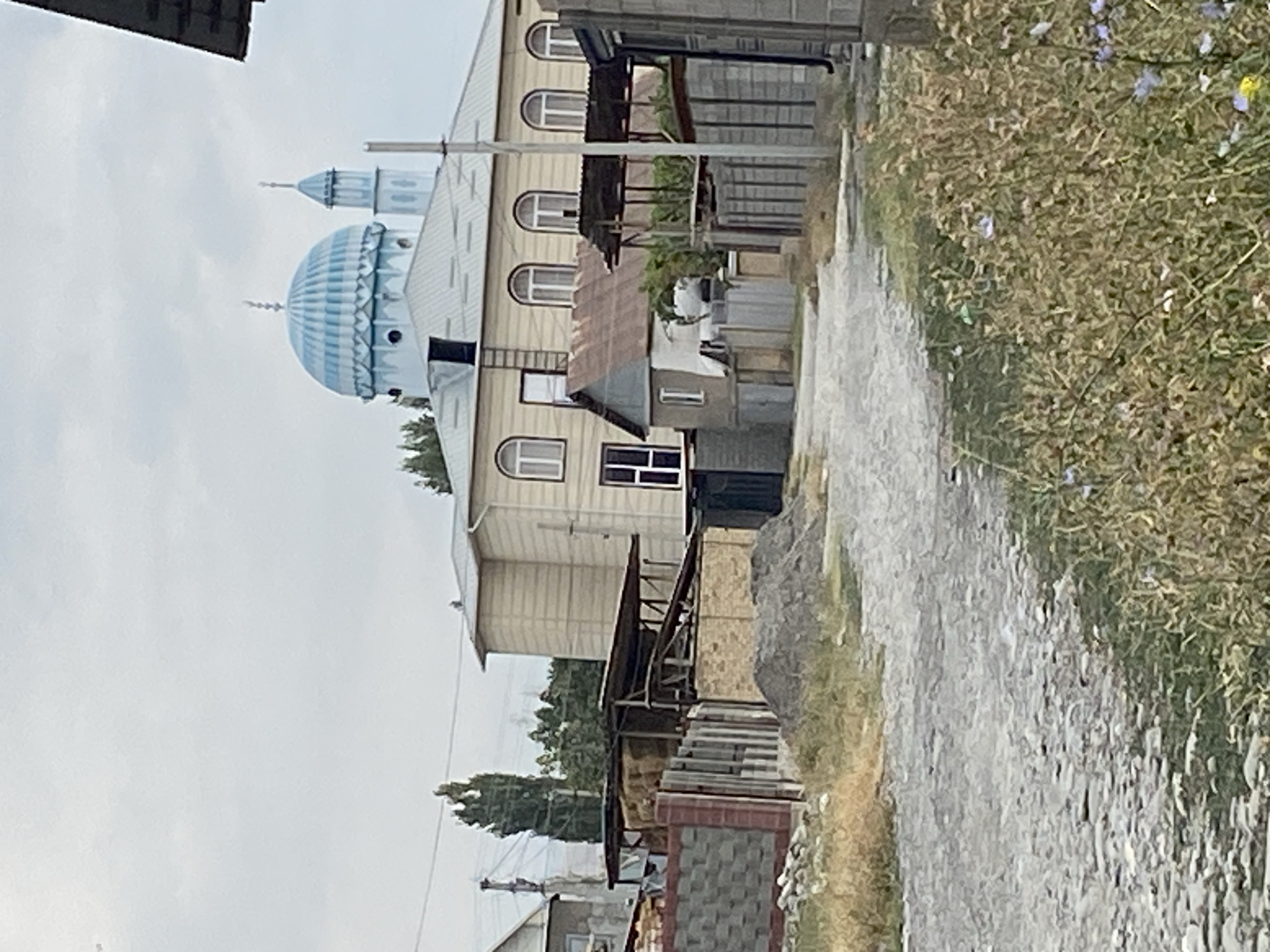 The image depicts a street scene in the city of Тайгараев, Kyrgyzstan, on an overcast day. The foreground is dominated by a rough, gravel road with dry grass and some small wildflowers growing along its edges. To the right, a wooden fence with vertical slats borders the road. Beyond the fence, a building with grey concrete blocks is visible.
In the middle ground, a tan-colored building with multiple arched windows and a light-colored metal roof stands prominently. A distinct blue-domed mosque with a slender minaret is situated behind and to the left of this building, its blue dome contrasting with the muted tones of the surroundings. Next to the tan building, a smaller, dilapidated structure with a rusty metal roof is present. Further to the left, there is a covered area filled with hay bales, bordered by a decorative stone and brick wall.
The background features trees and overhead power lines, suggesting a residential or semi-rural area. The sky is uniformly grey and cloudy. There are no people or animals visible in the image. The overall impression is one of a quiet, possibly developing, neighborhood.