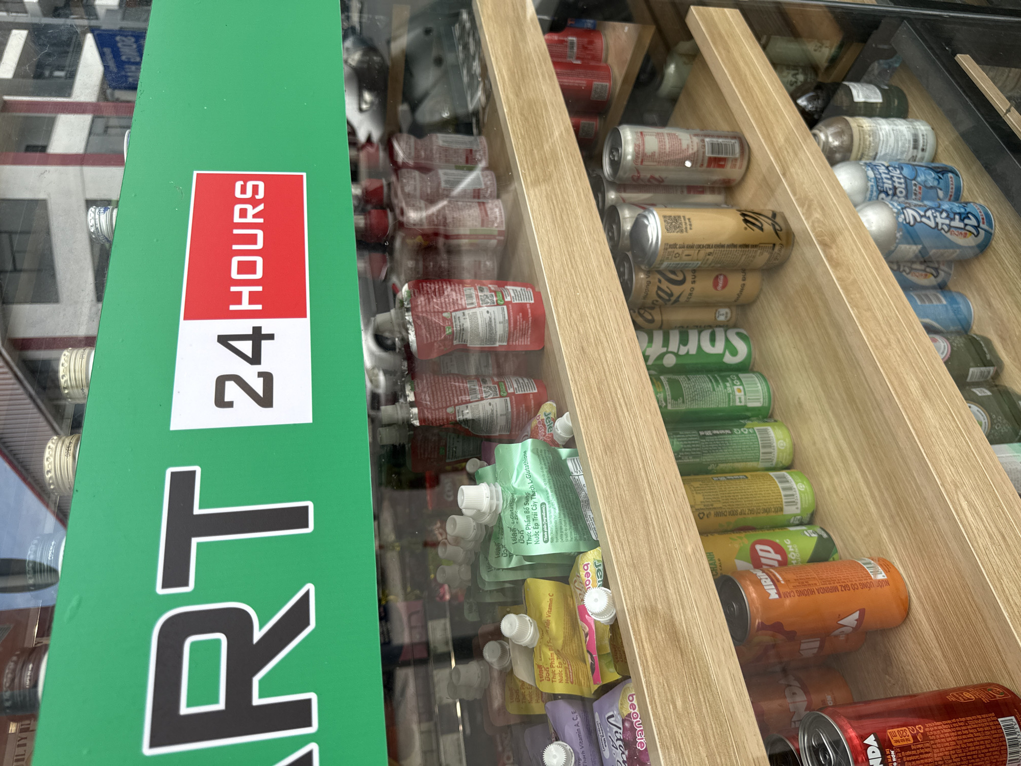 The image shows the front of a small convenience store, likely a 24-hour Mart, in Nha Trang, Vietnam. A bright green banner across the top of the display window reads "ART 24 HOURS". Inside the refrigerated display, shelves are stocked with various beverages. The top shelf holds pouches of juice or similar drinks, some with Vietnamese text. The second shelf is filled with canned drinks, including Sprite and Coca-Cola, along with other brands. The bottom shelf contains more canned drinks and some bottled beverages. The overall impression is of a well-stocked convenience store aiming to attract customers with its round-the-clock availability.