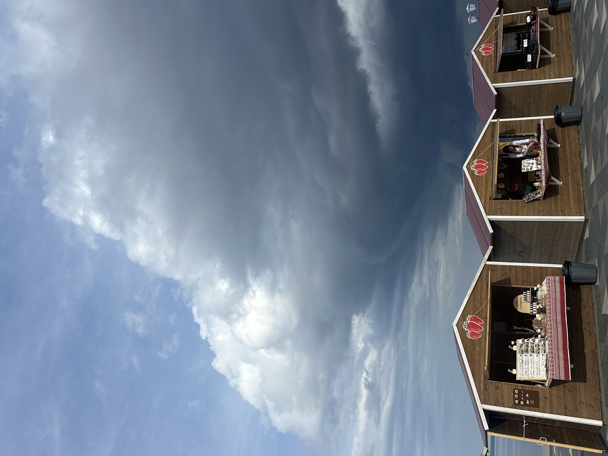The image shows a row of three wooden market stalls with red roofs, positioned along a paved waterfront promenade. The stalls are displaying various goods, including jewelry and what appear to be bottles of wine or other beverages. The sky above is dramatic, with dark, heavy storm clouds gathering and moving in from the right, contrasting with lighter, wispy clouds and patches of blue sky on the left. The scene is located in Petrozavodsk, Russia, likely on the shore of Lake Onega, given the presence of water on the left side of the frame and the characteristic landscape often associated with the city's embankment. A single woman is visible inside one of the stalls, attending to customers or arranging merchandise. The overall atmosphere is one of impending weather change, with the natural elements of the sky creating a striking backdrop for the commercial activity.