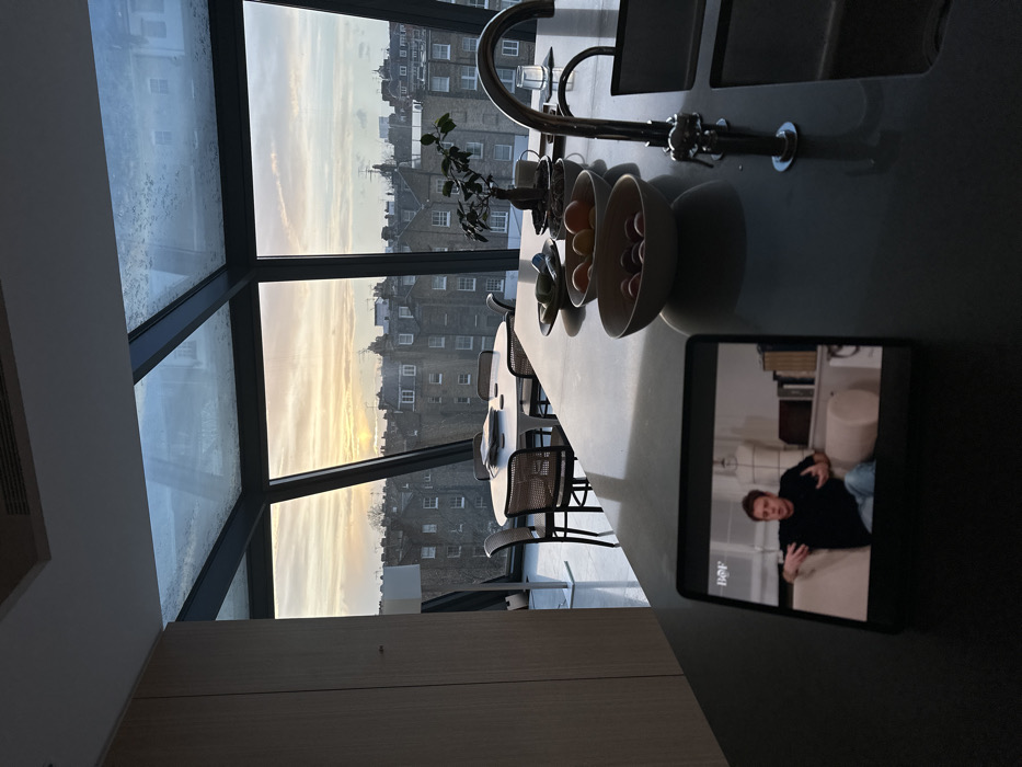 The image displays a modern interior space, likely a kitchen or dining area, with a panoramic view of London's urban landscape through a large, angled window. The window structure suggests a conservatory or a very tall, contemporary design, offering abundant natural light.
On a long, light-colored counter, a chrome kitchen faucet is visible, alongside a small potted plant. Bowls containing fruit, such as oranges or tangerines, and potentially eggs, are also arranged on the counter. Wicker-backed dining chairs are pulled up to a section of the counter, indicating a dining space.
A tablet or similar device rests on the counter, displaying a video. The screen shows a young man indoors, dressed in a dark top and light pants, making a hand gesture with both hands. The word "Bof" is visible in white text on the lower left of the screen.
The time of day appears to be either sunrise or sunset, with the sky showcasing soft hues of yellow, orange, and pale blue on the horizon, illuminating the rows of distinctive London buildings. Some condensation or raindrops are visible on the upper window panes, hinting at cool weather or recent precipitation. The scene conveys a sense of calm and modern urban living in London, United Kingdom.