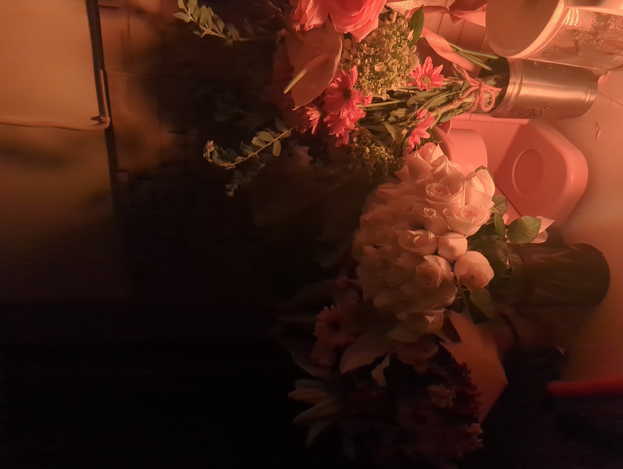 The image shows a floral arrangement in a pink container inside what appears to be a cupboard. The arrangement includes roses, anthuriums, and daisies, along with greenery. A metal thermos is also visible in the arrangement. The scene is located in Kediri, Indonesia.