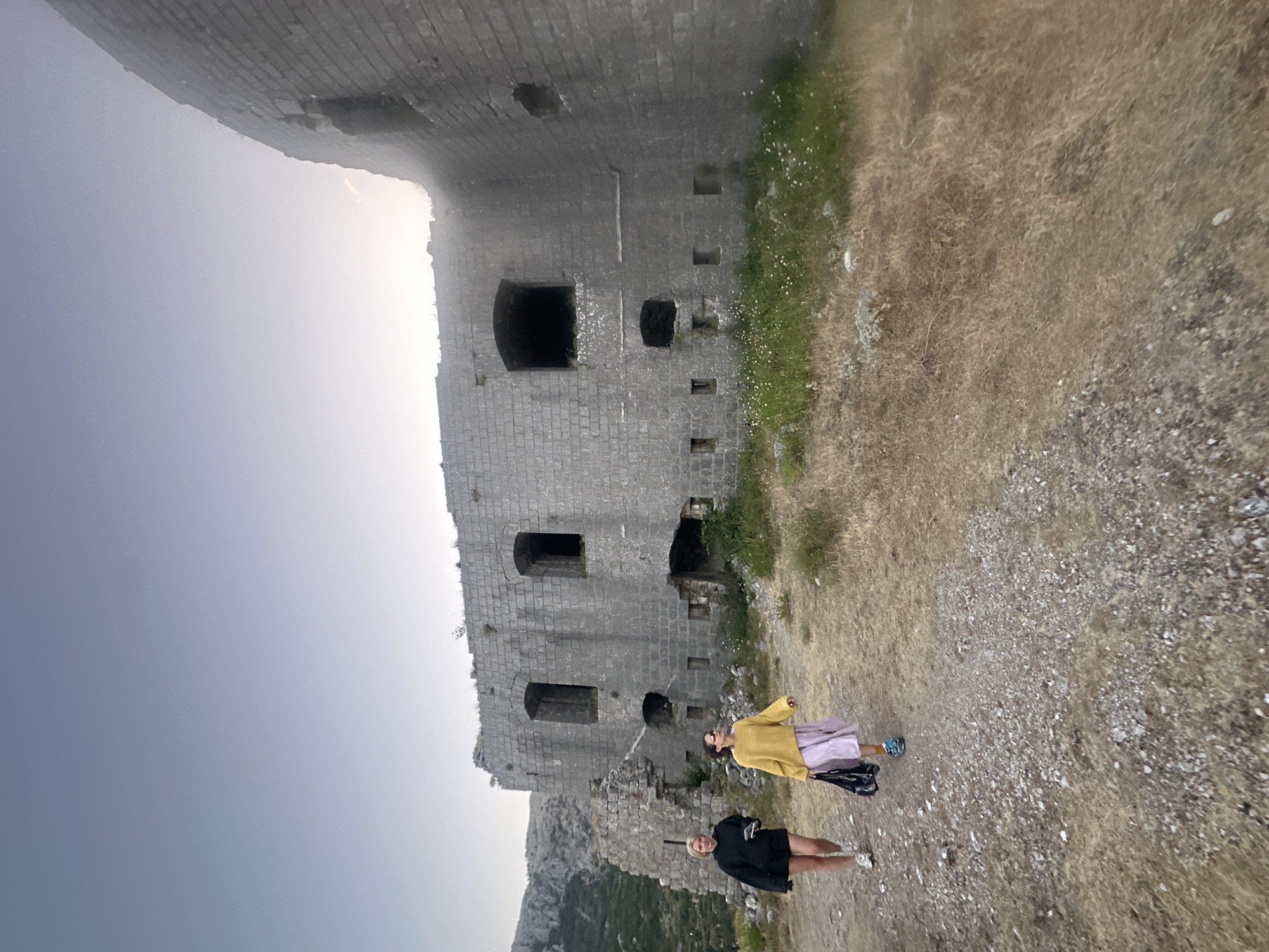 Two individuals walk on a gravel path towards a weathered, stone fortress ruin in Brajici, Montenegro. The person on the left, with blonde hair, wears a black hooded sweatshirt and black shorts, carrying a phone in their left hand. The person on the right, with dark hair, is dressed in a bright yellow sweater and a light purple skirt, holding a dark bag. The fortress displays crumbling stonework and arched openings, indicative of age and disrepair. The surrounding landscape consists of dry grass, scattered small white flowers, and rocky terrain, with a mountainous backdrop under a hazy sky. The lighting suggests it is late afternoon or early evening.