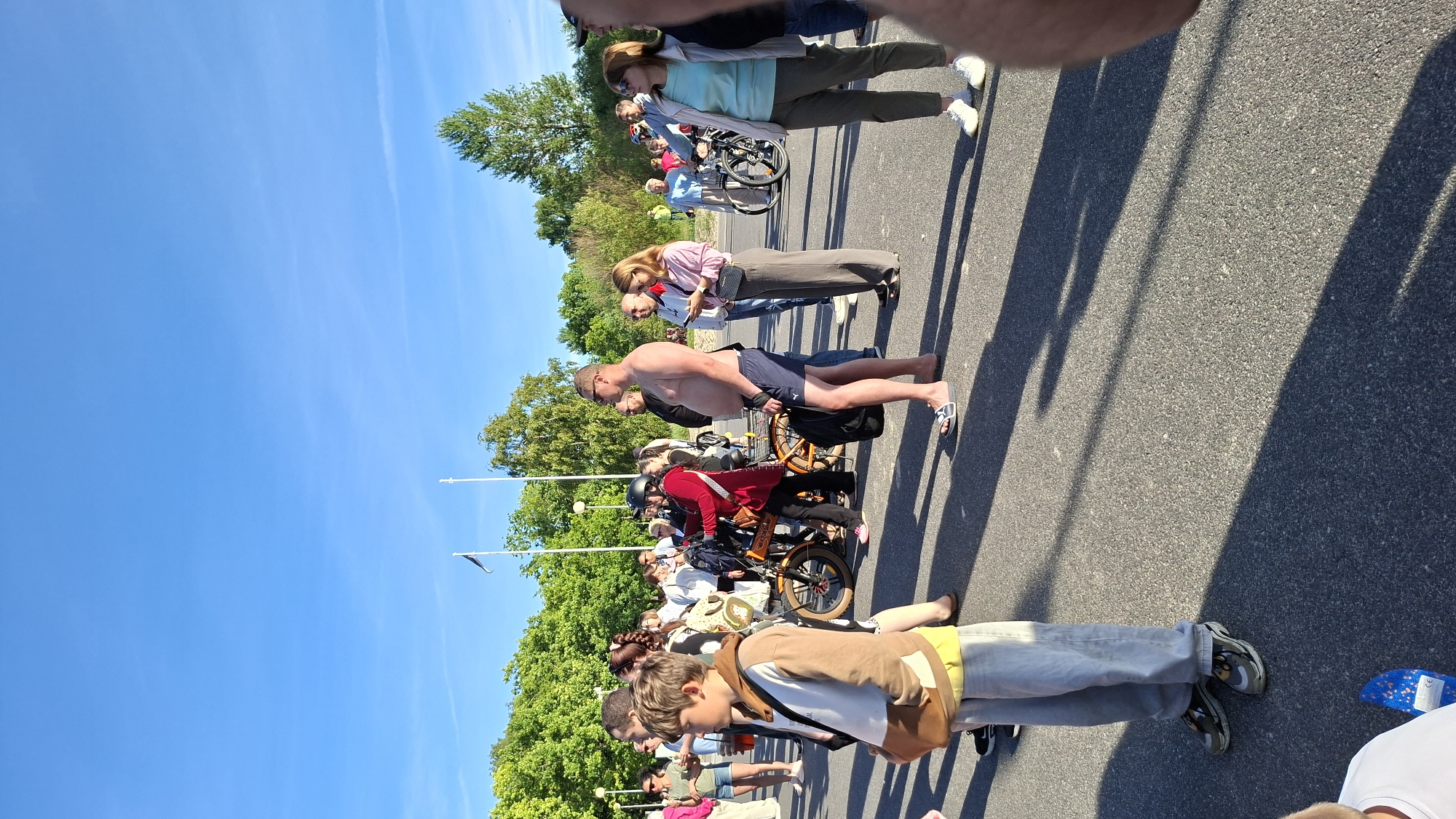 The image depicts an outdoor scene with a group of people standing on an asphalt surface, likely a street or parking lot, with a clear blue sky overhead. The environment suggests a public space, possibly a park or event area, with trees visible in the background. Several individuals are gathered, some are looking in different directions, and others appear to be engaged in conversation. Bicycles are present, indicating a possible cycling event or recreational activity. The time of day is likely daytime, given the bright sunlight casting shadows. The attire of the people suggests a casual setting. The presence of flags in the background might indicate a civic event or a location with national symbolism. Given the context of this image, it is very likely taken in Tallinn, Estonia, a city known for its public spaces and cycling culture.