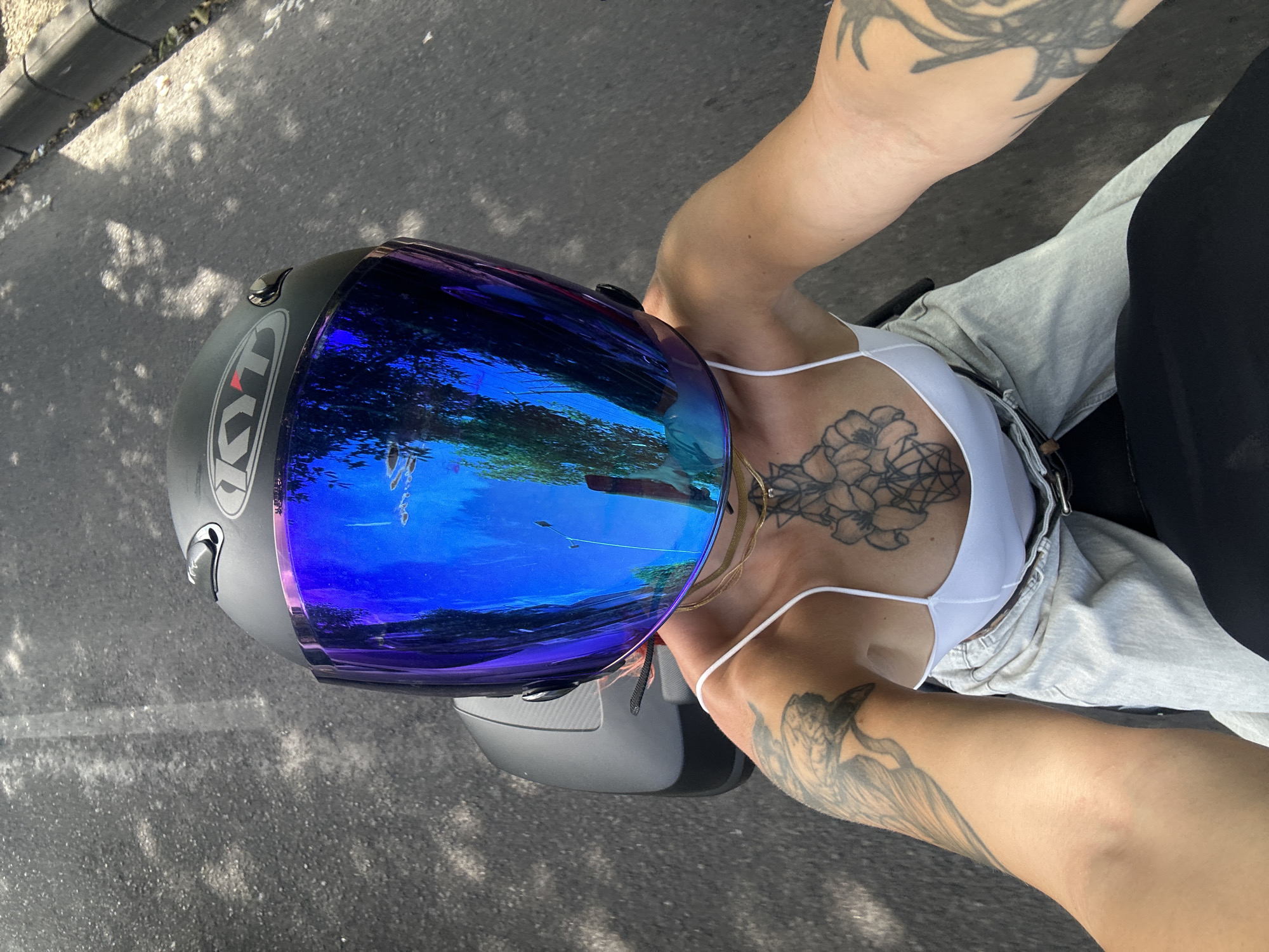 The image shows a person on a motorcycle from an overhead perspective, likely taken by someone on the same vehicle. The person is wearing a grey KYT helmet with a blue mirrored visor. Their visible skin is light-toned and adorned with tattoos on both arms and a chest piece. They are wearing a white top and grey pants, with a glimpse of a black garment beside them. The motorcycle is on asphalt, probably a road. The reflection in the visor shows trees and sky, typical of a city environment. The location is consistent with Kuta, Indonesia, given the likely weather, vegetation, and clothing style.