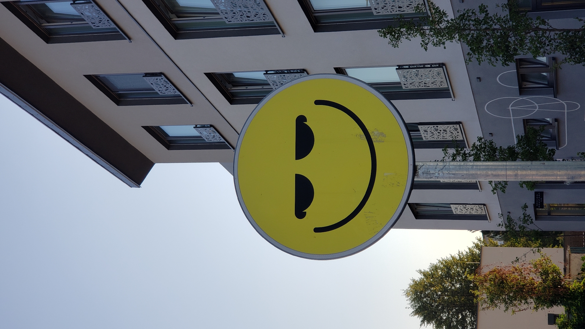 The image shows a yellow, oval-shaped sign with a smiley face on it. The smiley face has thin, horizontal, closed eyes and a wide, curved smile. The sign is mounted on a silver pole, which is partially visible. In the background, there is a modern, multi-story building with light-colored walls and dark-framed windows. Some of the windows have decorative balcony railings. Trees and vegetation are visible in front of and around the building. The sky is clear and light blue. The overall scene appears to be outdoors, possibly in an urban or suburban setting. There are no people visible in the image. The sign is prominently displayed and appears to be a form of public art or a decorative element. The location of Prešov, Slovakia is mentioned, but no specific landmarks or context within the city are provided in the image itself to confirm this location.