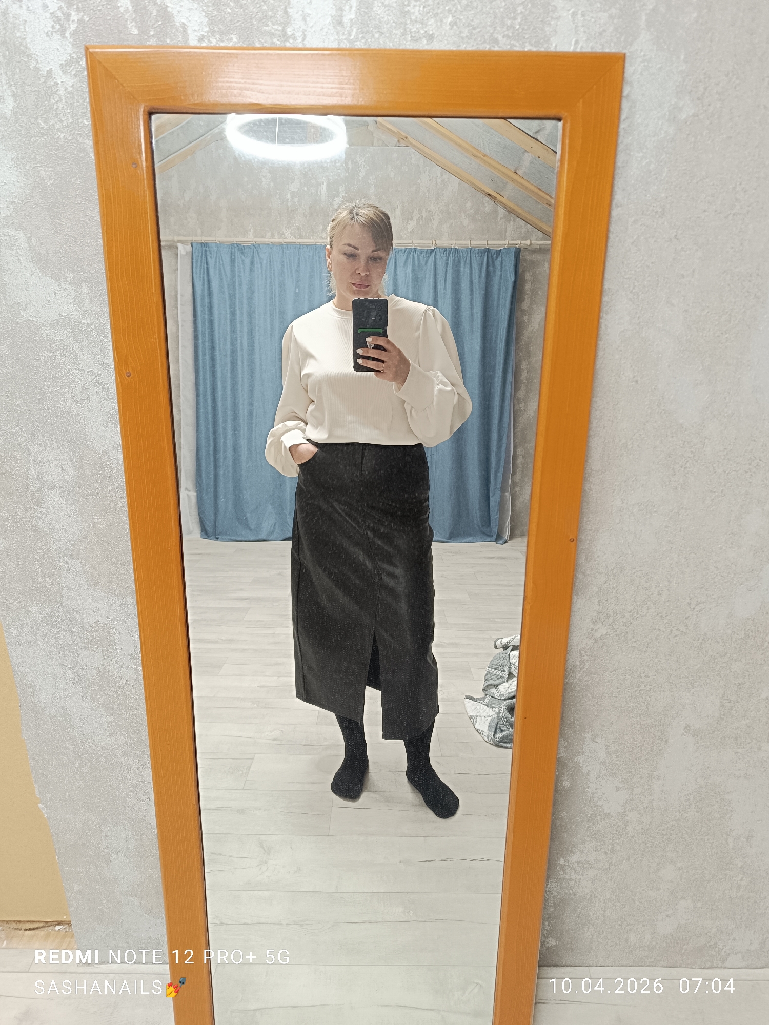 A woman is standing in front of an orange-framed mirror, taking a photo of herself with a smartphone. She is wearing a long-sleeved, white ribbed shirt with puffed sleeves and a long, dark skirt with a front slit. She is also wearing black socks and shoes. Her hair is pulled back in a bun. She has one hand in her pocket. The background shows a room with light blue curtains and an unfinished ceiling. The walls are painted a neutral, light color. The room is located in Chaikovsky, Russia, as the timestamp at the bottom of the image indicates the date and time are specific to that location.