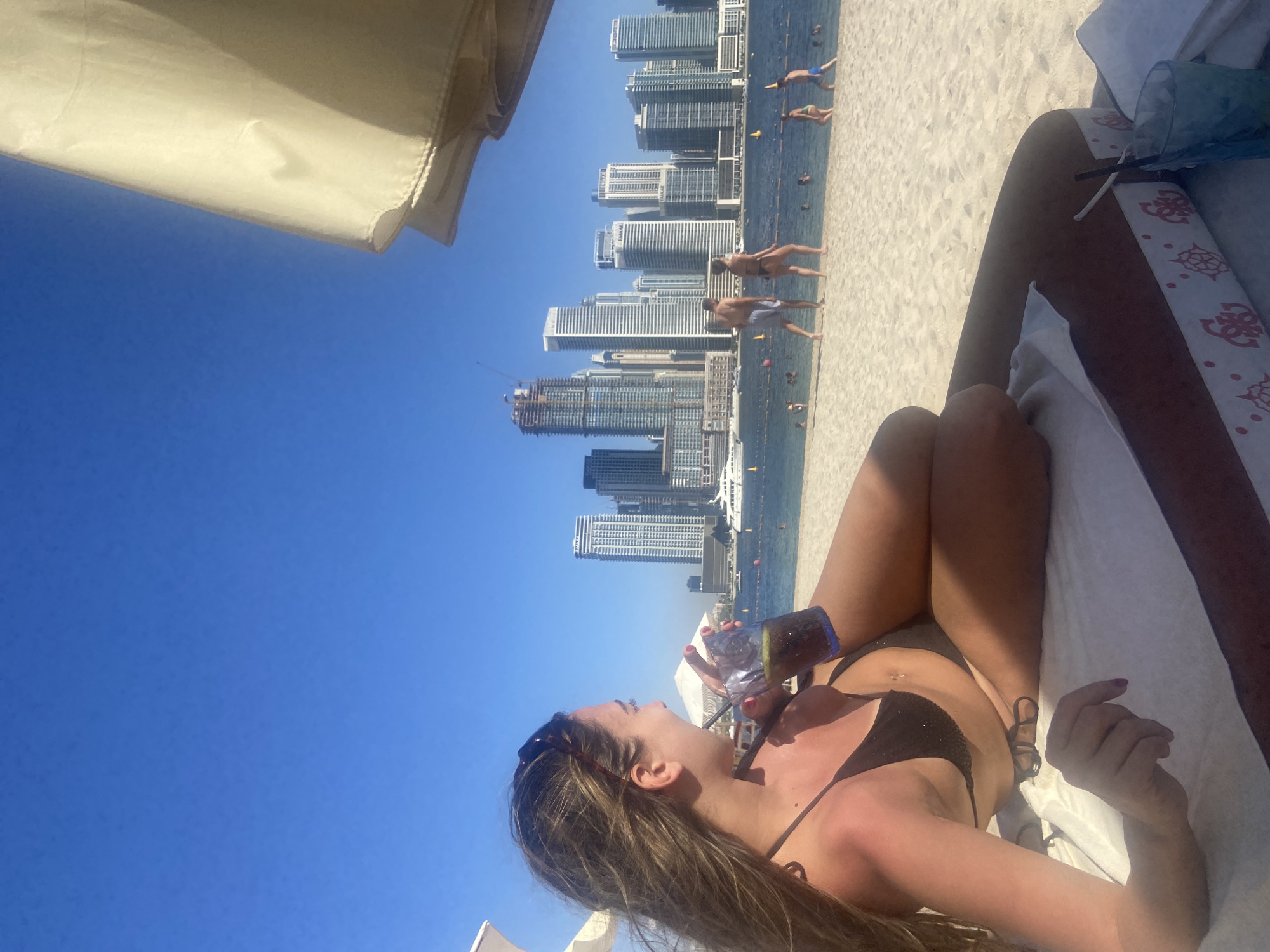 The scene depicts a woman in a black bikini lying on a white towel on a beach, holding a drink in a blue glass. In the background, there is a coastline and cityscape, with multiple high-rise buildings identifiable as being in Dubai, UAE. People are visible walking and swimming in the water near the shore. A beach umbrella is partially visible in the top left of the frame.