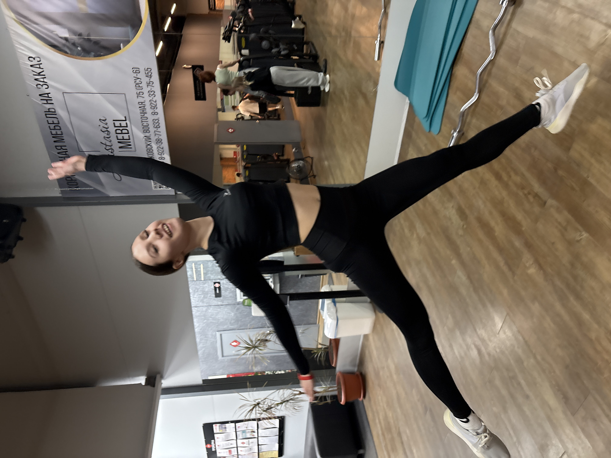 An indoor full-shot captures a young woman performing a side-splits or stretching pose in what appears to be a gym or fitness studio in Chaikovsky, Russia. She has dark hair tied back and is wearing a black long-sleeved crop top, black leggings, white socks, and light-colored athletic shoes. She stands on her left leg, extending her right leg outward, with her left arm reaching down and her right arm extending upwards, balancing herself. She is smiling towards the camera, conveying a sense of happiness and engagement.
The background shows a wooden floor and various gym equipment, including several weight machines, where other indistinct figures are visible, possibly also exercising. A bright blue yoga mat and a silver barbell are resting on the floor to the woman's right. Potted plants and a white trash receptacle are also visible. A large banner on the wall serves as a prominent background element. The banner features Russian text, which reads: "КОРПУСНАЯ МЕБЕЛЬ НА ЗАКАЗ" (Custom cabinet furniture), "Anastasia MEBEL" (Anastasia Furniture), and an address "ЧАЙКОВСКИЙ, ВОСТОЧНАЯ, 75 (РСУ-6)" (Chaikovsky, Vostochnaya, 75 (RSU-6)), along with phone numbers "8-922-38-77-633, 8-922-33-75-455". The scene is brightly lit by overhead lights, suggesting daytime.