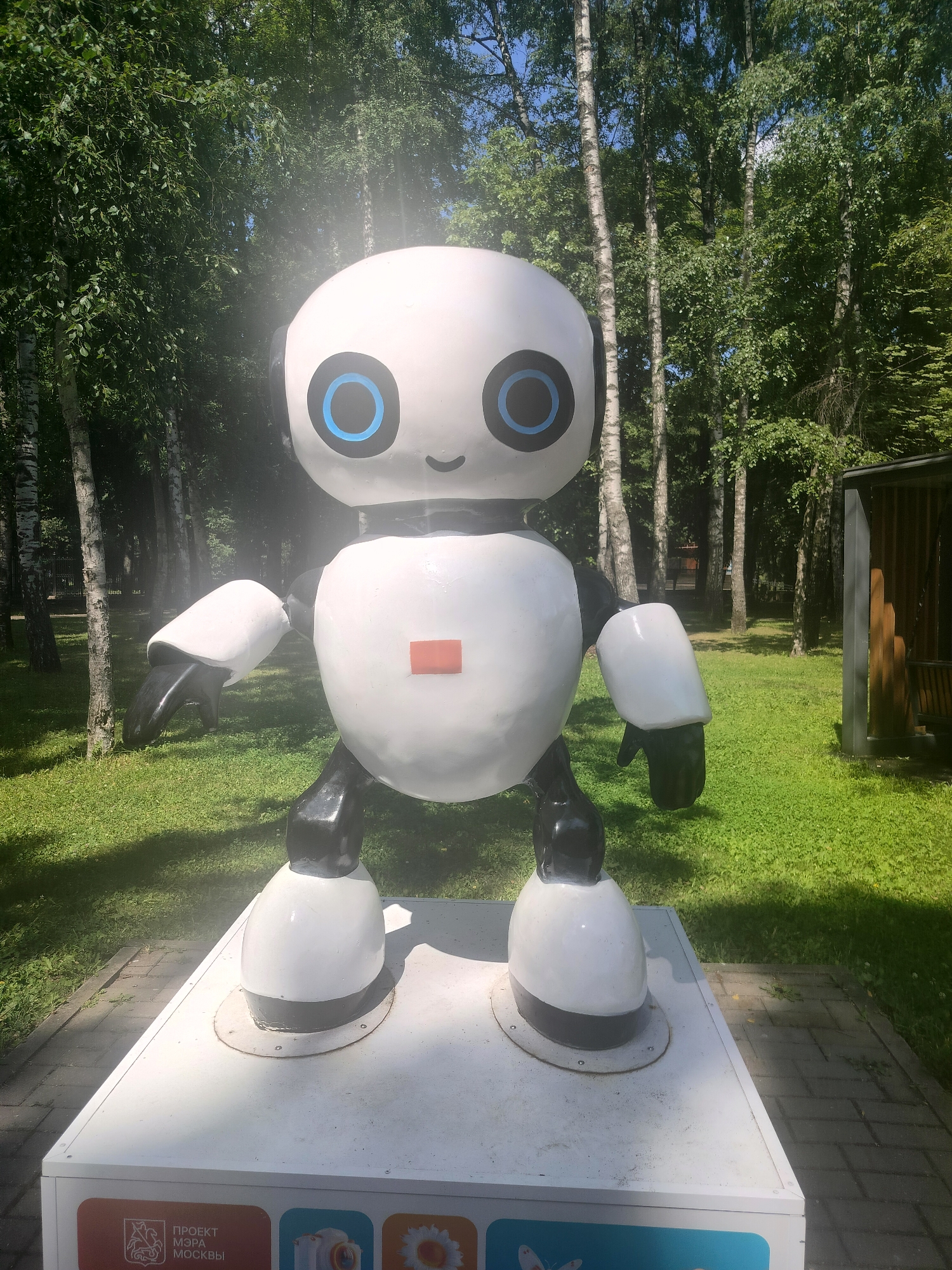 A life-sized, white and black robot sculpture stands on a white pedestal in a park setting. The robot has a round head with large blue circles for eyes and a small, smiling mouth. A small red rectangle is centered on its chest. Its arms and legs are segmented and black at the joints, with white outer shells. The robot is positioned in front of a line of birch trees with lush green foliage. The ground is covered in green grass, and a stone pathway is visible to the right. A wooden structure, possibly a gazebo, is partially visible in the background on the right. The base of the pedestal has an inscription in Russian, which includes "ПРОЕКТ МЭРА МОСКВЫ" (Mayor's Project of Moscow). The scene appears to be outdoors on a sunny day, with dappled sunlight filtering through the trees.