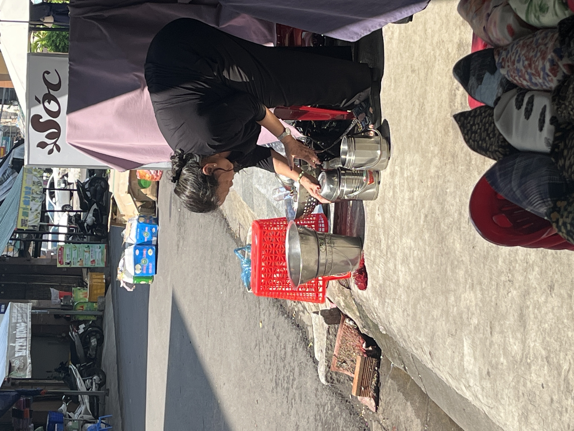A medium shot displays a street vendor in Nha Trang Ward, Vietnam, operating a food stall on a sunny day. The vendor, dressed in black, appears to be preparing or serving food, interacting with stainless steel containers placed on a surface behind a red plastic basket. The background includes parked motorcycles and buildings. A stall with various hats and possibly other goods is visible on the right. The scene is a typical urban setting with commercial activity.