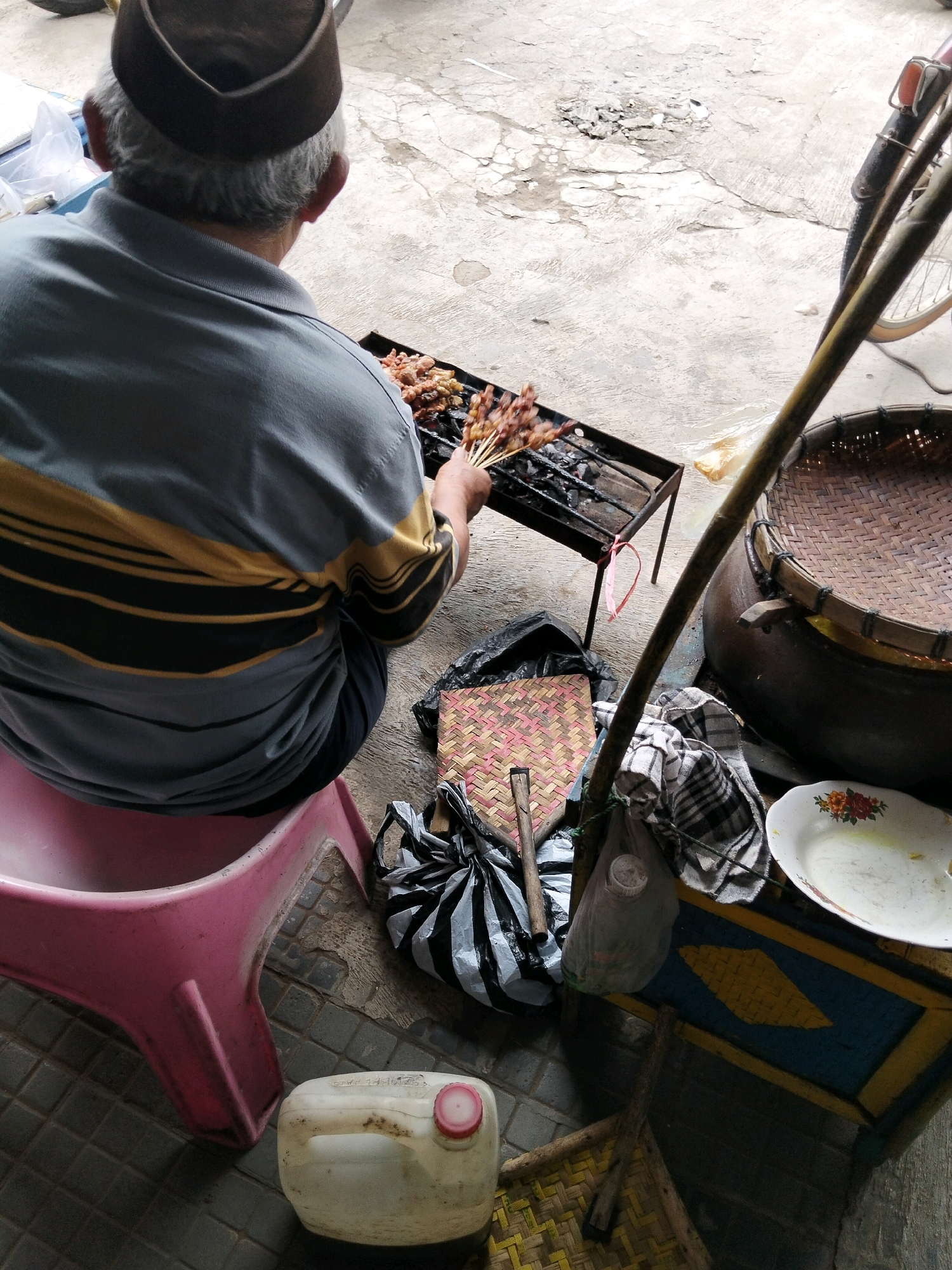In Kemiri Kidul, Indonesia, a person wearing a brown cap and a grey and yellow striped shirt sits on a pink stool, tending to satay on a charcoal grill. The grill is positioned on a metal stand. In the background, a bicycle is partially visible, along with a large woven basket. Various items are scattered around the scene, including plastic bags, a woven mat, a plastic jug, and a plate. The ground is paved with concrete and stone tiles.