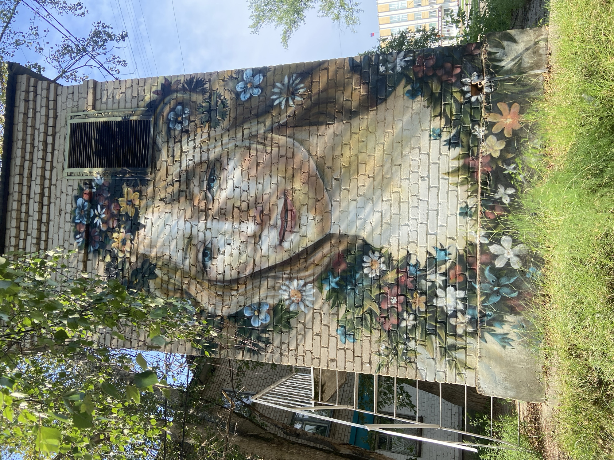 A mural of a woman's face with blue eyes and blonde hair is painted on a brick building. She is surrounded by flowers and leaves. The building has a metal ventilation grate on the side. There is a white metal staircase on the left side of the building. The background shows trees and a building in the distance. This mural is located on an electrical substation at 46 Vokzalnaya Street, Chaikovsky, Russia.