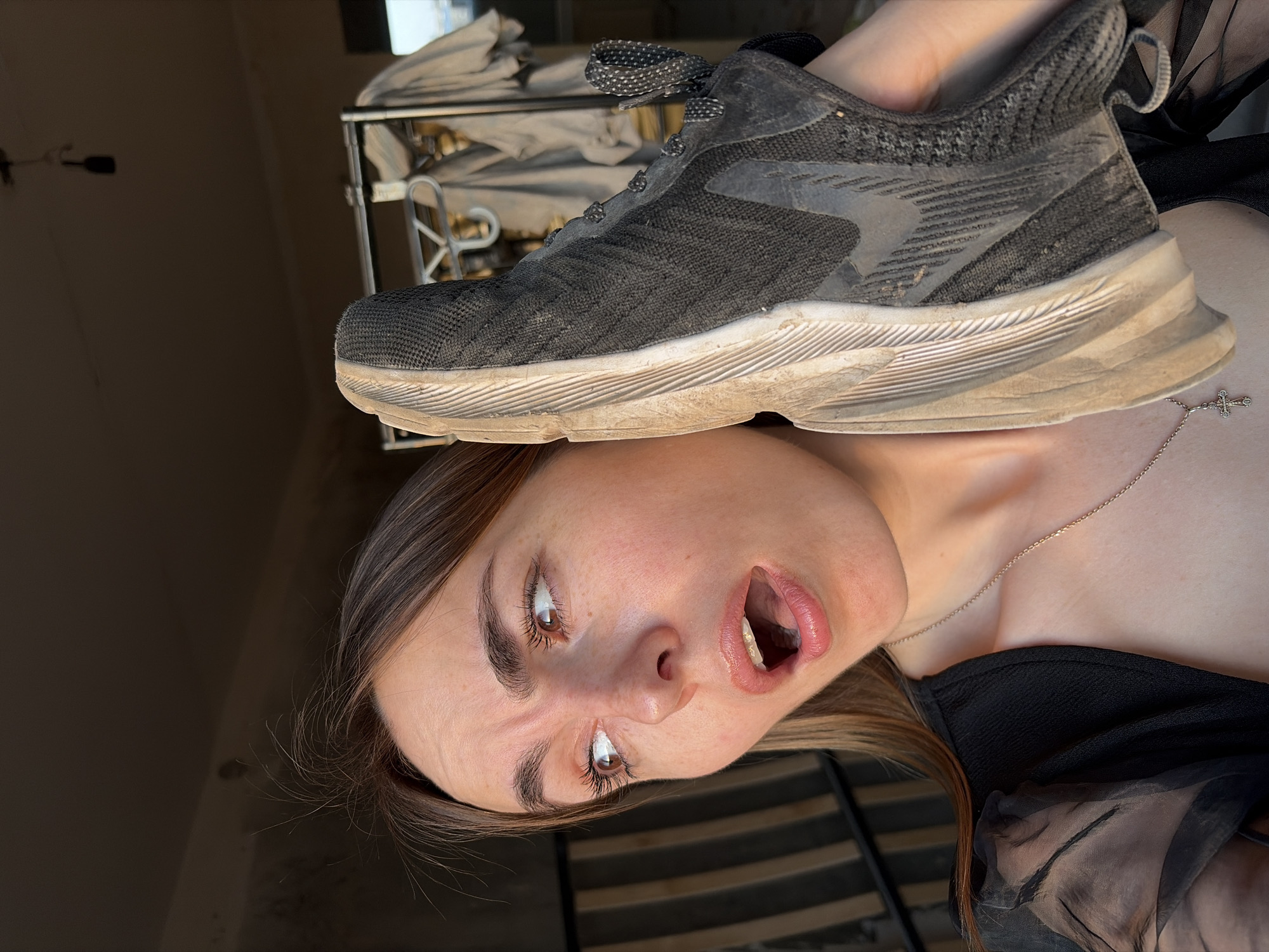 A woman lies on her back with a dirty athletic shoe placed on her forehead. The woman has fair skin, light brown hair, and is wearing a black top with sheer sleeves. A silver necklace with a cross pendant is visible on her chest. Her mouth is open, and she appears to be looking up and to her left. The shoe is dark gray with a white sole and gray laces. It is visibly dirty, with smudges and grime on the sole and upper.
In the background, a partially visible metal rack with light-colored items draped over it is present. The background also reveals a wall and part of a slatted surface, possibly a bed frame or similar structure.
There is no information about the city of Izhevsk, Russia in this image.