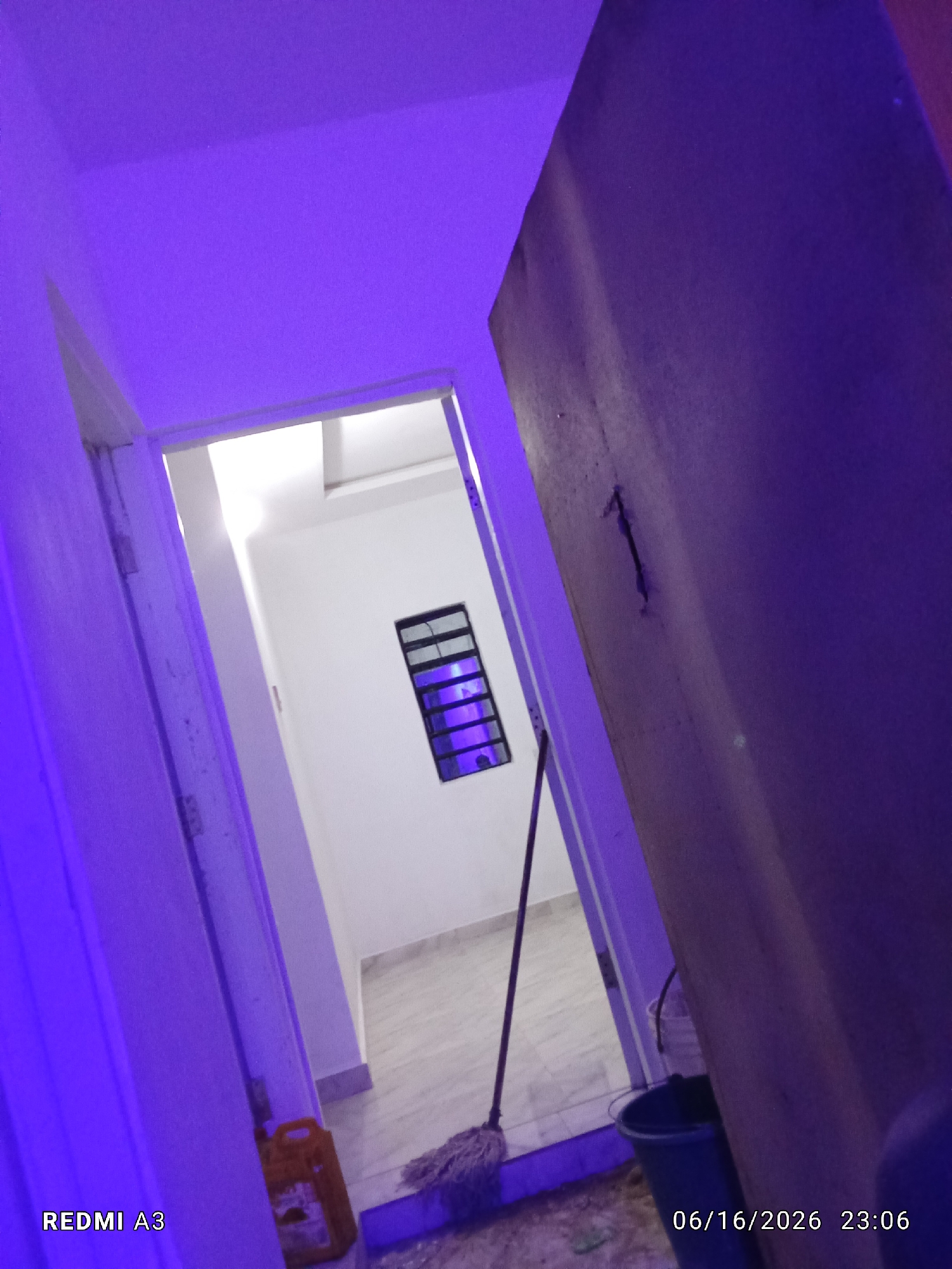 The image shows an interior scene, possibly a hallway or entryway. The walls and ceiling are illuminated with a strong purple light, creating a dramatic and somewhat surreal atmosphere. To the left, a doorframe is visible, painted white and showing signs of wear. Through an open doorway ahead, a brightly lit room with white walls can be seen. A small, rectangular window with dark panes is mounted high on the far wall of this room. A mop with a light-colored head rests against the wall, with its handle extending upwards and out of frame. To the right of the doorway, a large, dark-colored door dominates the foreground, obscuring much of the right side of the image. A yellow plastic container is visible on the floor near the doorway, and a dark blue bucket stands near the large door. The floor appears to be tiled. The timestamp in the bottom right corner indicates the date as 06/16/2026 and the time as 23:06. The text "REDMI A3" is visible in the bottom left corner. The overall impression is one of a functional space, perhaps a home or business, illuminated by colored lighting. There are no people visible in the image.