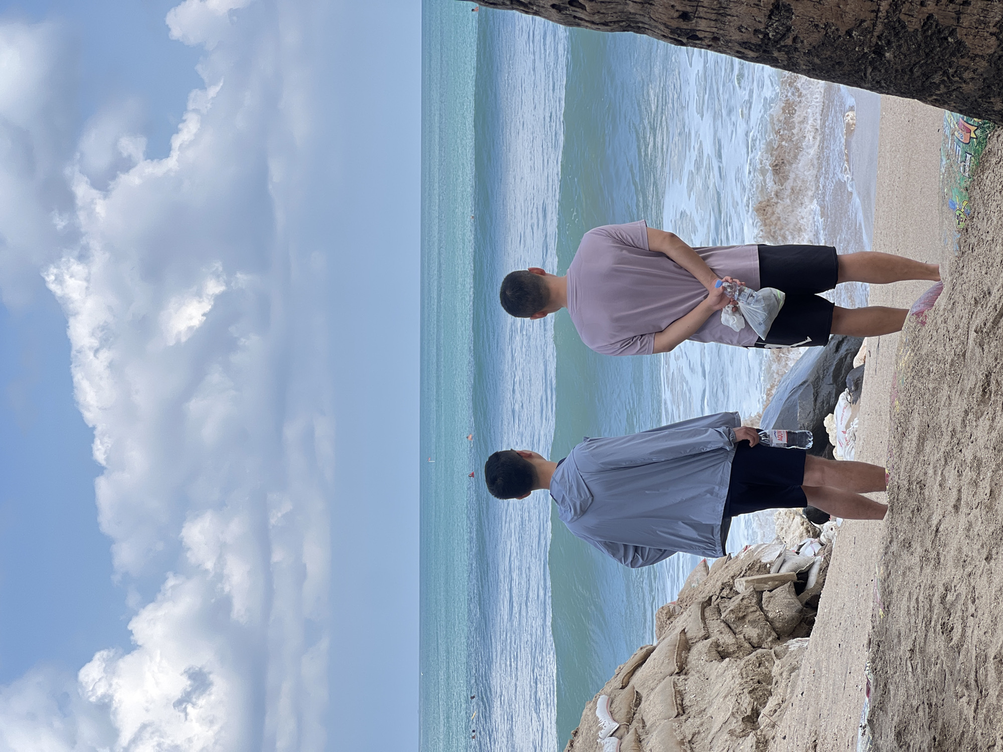 The image depicts a beach scene in Kuta, Indonesia, with two men standing on the sand facing the ocean. The man on the left wears a grey hooded jacket and black shorts, holding a bottle of Aquvit water. The man on the right wears a mauve t-shirt and black shorts, holding a plastic bag in his clasped hands. In the background, the turquoise ocean has moderate waves, and several people are visible swimming and surfing. The beach area is covered in sand and debris, with sandbags and rocks forming a barrier to the left. A portion of a palm tree trunk is visible on the right side of the frame. The sky is partly cloudy with a light blue hue.