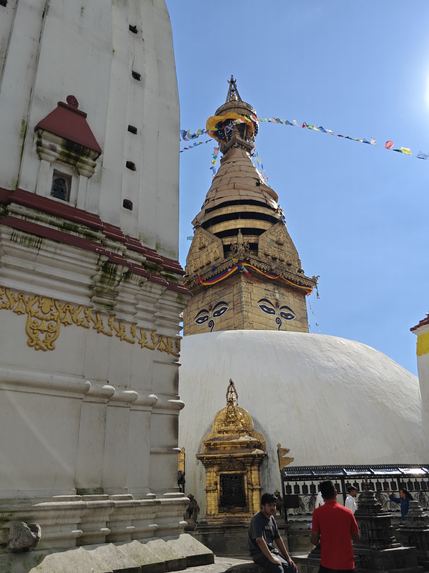 The image shows the Swayambhunath Stupa, a UNESCO World Heritage Site located in the Kathmandu Metropolitan City, Nepal. The stupa is a prominent landmark, characterized by its golden spire adorned with prayer flags and painted eyes, symbolizing wisdom and compassion. In the foreground, a large white stupa structure is visible to the left, with intricate golden detailing and tiered roofs. Several people are present, including a man in a red shirt and another in a grey t-shirt sitting on a ledge. Other individuals are seen in the background near prayer wheels. The sky is clear and blue, suggesting a sunny day. The architecture features a blend of white painted structures and ornate golden decorations, typical of Nepalese religious sites.