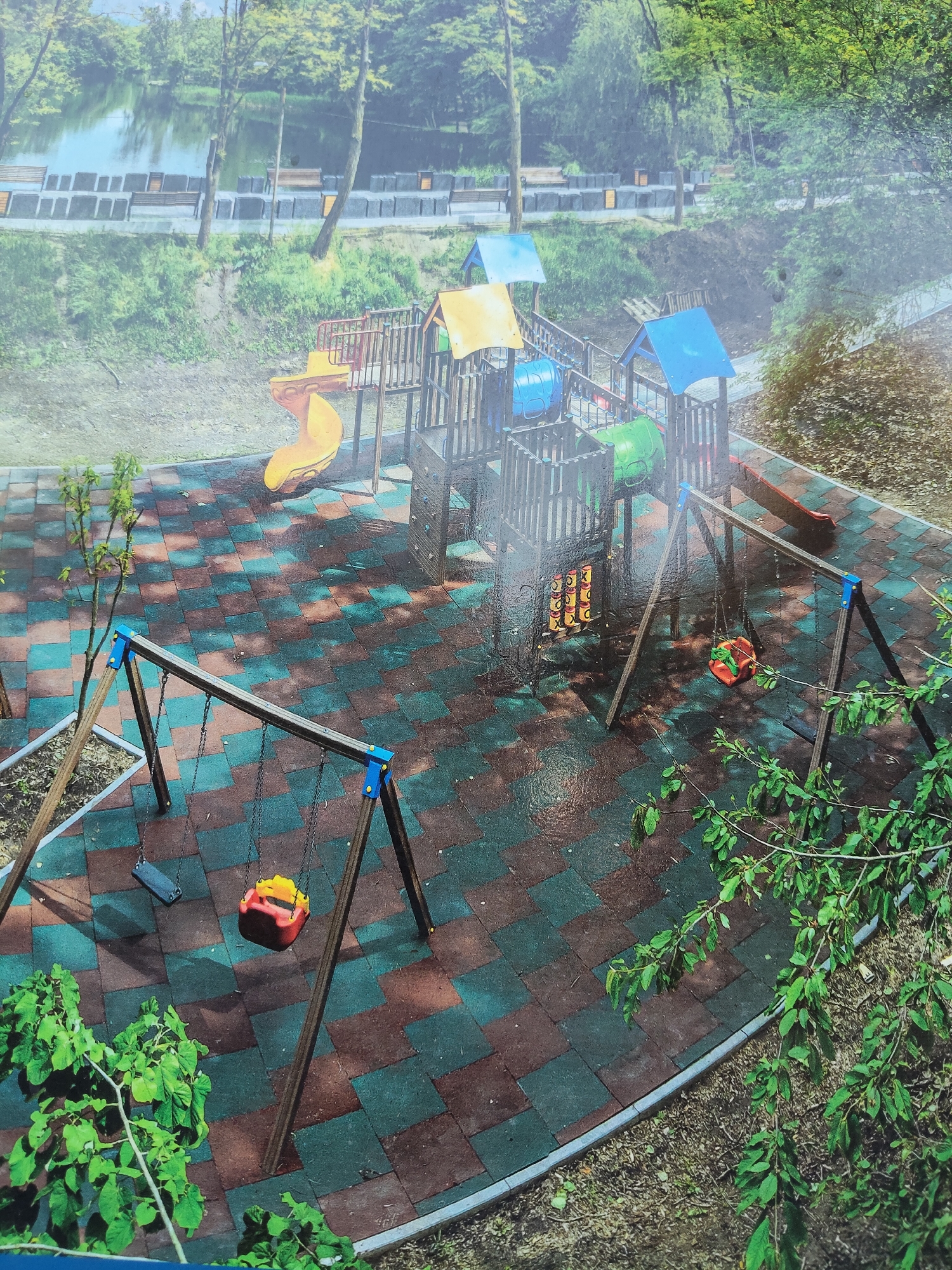 The image depicts a children's playground with a colorful, modular play structure featuring slides, tunnels, and climbing elements. The ground is covered in a rubberized surface composed of interlocking square tiles in shades of red-brown and teal. Two swing sets are visible, each with two swings. One swing set has a single red and yellow swing, and another has a single blue swing. The playground is situated near a body of water, possibly a lake or river, with trees and greenery surrounding it. In the background, there are benches and what appear to be tiered structures along the water's edge. The overall impression is of an outdoor recreational area that appears to have recently been rained on, as evidenced by the wet surfaces. The scene does not contain any people. The location is a children's playground in Chișinău, Moldova.