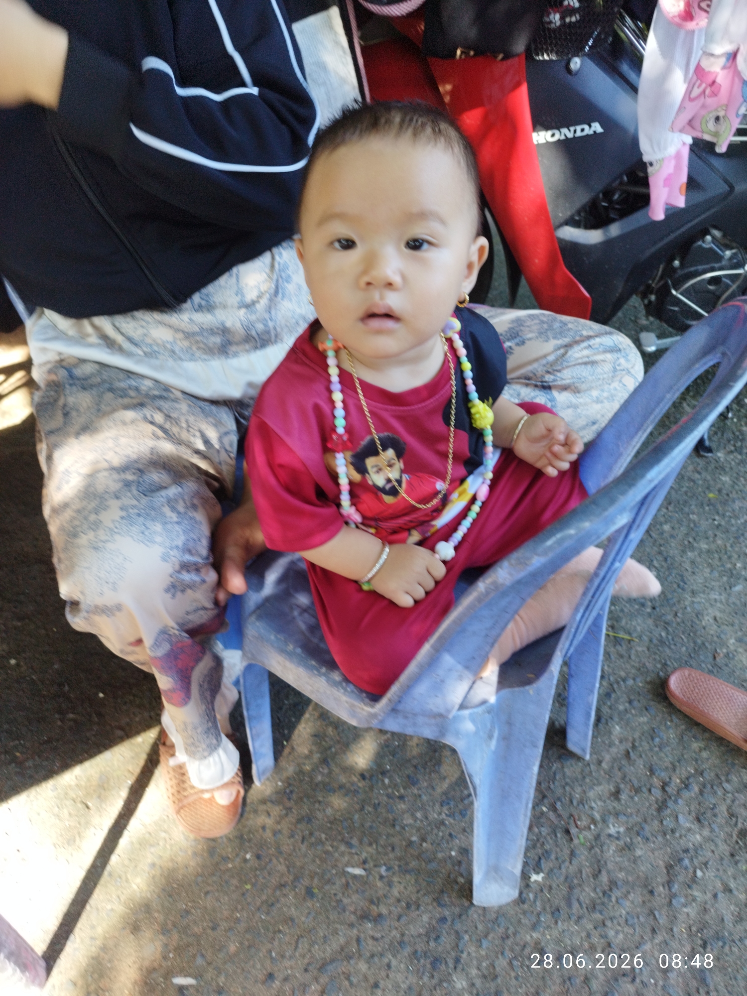 A young child wearing a red shirt with a picture of Mohamed Salah on it and a colorful beaded necklace sits on a blue plastic chair. The child is wearing a gold earring and beaded bracelets on each wrist. The child's legs are crossed, and they are wearing brown woven shoes. The child is sitting on a person's lap, who is wearing patterned pants. In the background, a motorcycle is partially visible, with the word "HONDA" on it. The scene is outdoors, on a concrete surface. A timestamp in the bottom right corner reads "28.06.2026 08:48". The image appears to have been taken in Mũi Né, Vietnam, likely on a street or sidewalk given the concrete ground and the presence of the motorcycle.