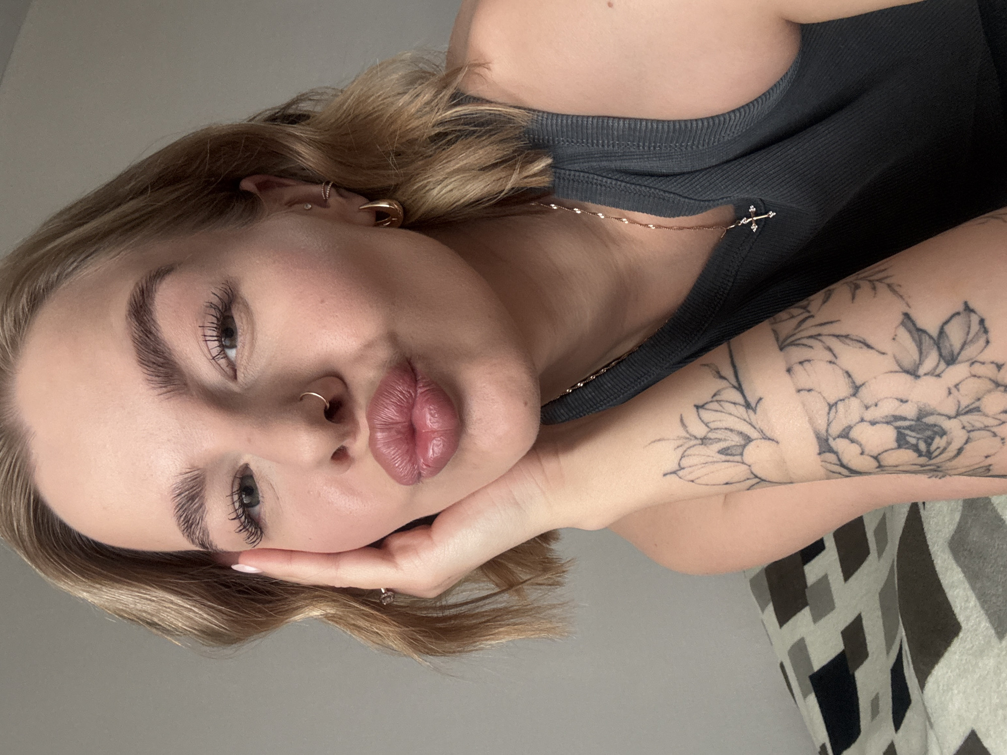 A young woman with blonde hair is posing for a selfie. She is wearing a gray tank top and a gold necklace with a cross pendant. She has a nose ring and earrings. Her arm is resting on a surface covered with a patterned textile. The background is a plain light gray wall. There is no information to indicate that this image was taken in Chaikovsky, Russia.