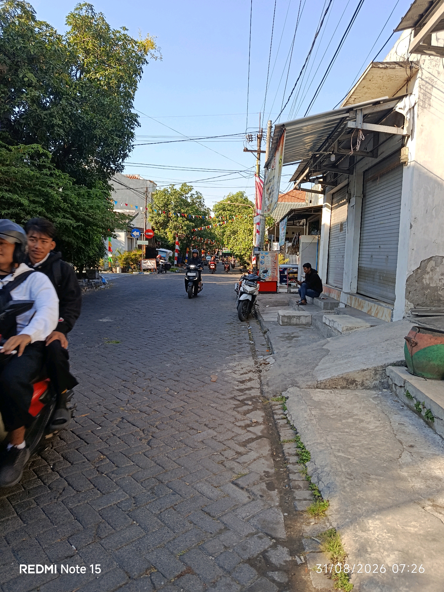 This image depicts a street scene in Keboansikep, Indonesia, captured on August 31, 2026, at 7:26 AM. The street is paved with dark grey cobblestones. On the left side of the street, two individuals are riding on a red scooter. The rider in front is wearing a helmet and a white top, while the passenger behind wears a black jacket. Further down the street, several other motorbikes are visible, with riders on some of them.
The right side of the street features a row of shops with roll-up metal doors, some of which are closed. An awning made of corrugated metal extends over the shop fronts. A man is seated on the concrete steps in front of one of the shops, looking down. A large green and orange cylindrical object, possibly a bin, is visible on the right edge of the frame. Trees with lush green foliage line both sides of the street. Colorful triangular flags are strung across the street in the distance, suggesting a festive occasion. Overhead, a network of power lines stretches across the sky, supported by utility poles. The sky is clear and blue, indicating a sunny morning. The overall impression is a typical morning street scene in a moderately busy urban area in Indonesia.
