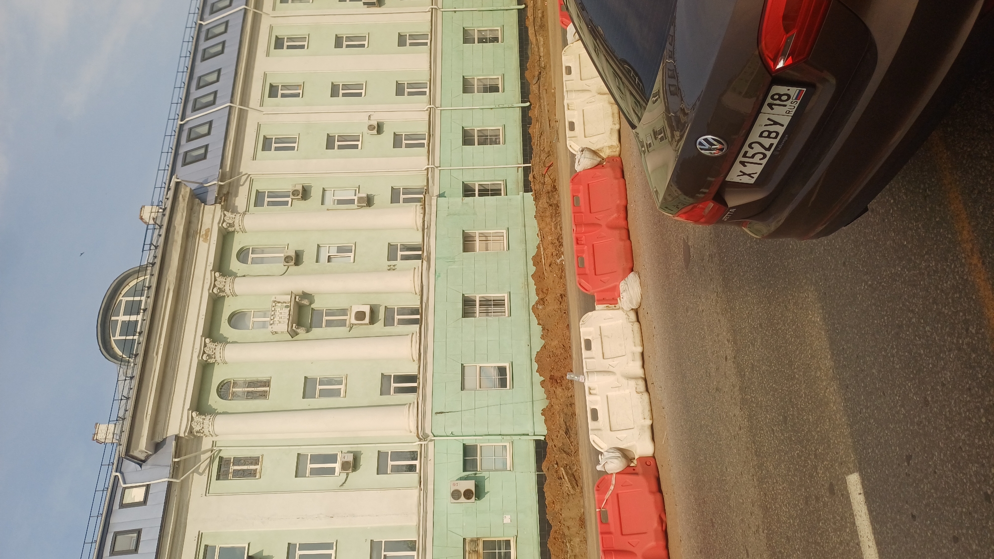 The image shows a view of a pale green building with white columns and arched windows, likely an administrative or institutional building. In front of the building, there is a construction site with piles of dirt and orange and white plastic barriers. A dark brown Volkswagen Jetta is parked on the right side of the frame, partially obscuring the construction barriers. The car's license plate reads "X152BY 18 RUS". The sky is blue with a few wispy clouds. The building has a mansard roof with dormer windows. The overall scene suggests ongoing construction or roadwork in a city environment.