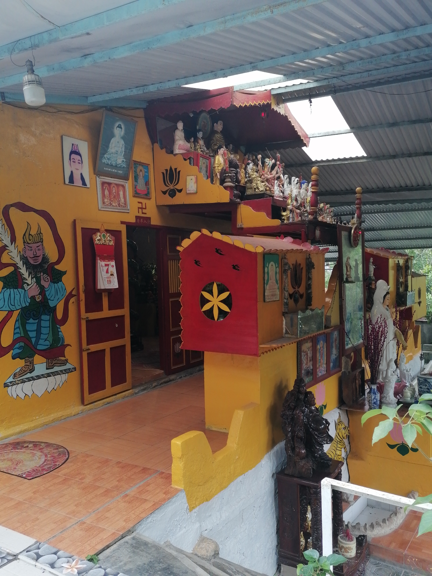 This image depicts a vibrant, richly decorated outdoor or semi-outdoor religious shrine in Phường Xuân Hương - Đà Lạt, Vietnam. The scene is brightly lit by natural daylight filtering through a corrugated metal roof, which features translucent panels.
The primary subject is a building with strikingly yellow walls and contrasting red doors and architectural elements. On the left wall, a large, colorful mural portrays a fierce guardian deity, possibly a Vajrapani, holding a jagged sword and standing on a cloud-like base. Above and to the right of this mural, several framed pictures are displayed. One prominent picture features a serene Buddhist figure, likely Guanyin, with the text "NAM MÔ A DI ĐÀ PHẬT" visible on its frame. Another portrait depicts a male figure with a distinctive head covering. A red and gold calendar, showing the number "7" prominently, hangs on a red, partially open wooden door. Nearby, a red Buddhist swastika symbol (Manji) is painted on the yellow wall. Above the adjacent red door frame, a Chinese inscription, possibly an auspicious blessing like "平安入室" (peace enters the house), is visible.
The central and right portions of the image are dominated by tiered altar-like structures, painted in red and yellow, densely populated with numerous statues and figurines. These include various depictions of Buddhas, Bodhisattvas (such as a large white statue of Guanyin on the right), and other revered figures, crafted from different materials and in diverse poses. Lotus motifs and other decorative patterns are integrated into these structures. Several potted plants, vases, and an ornate dark wooden stand holding more statues contribute to the intricate display. The ground is covered with reddish-brown patterned tiles, with a colorful round rug near the mural. A modern hanging light fixture is visible on the left side, suspended from the roof. The setting evokes a strong sense of devotion and cultural richness.
