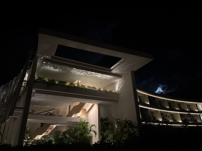 A modern building is shown at night. It features layered levels with open spaces and staircases. Green plants are visible on some levels. The design incorporates a rectangular opening on the top level. To the right is a curved building with illuminated balconies. The background is a dark sky with the moon peeking through the clouds. Shadows and light interplay create a visually interesting architectural scene.