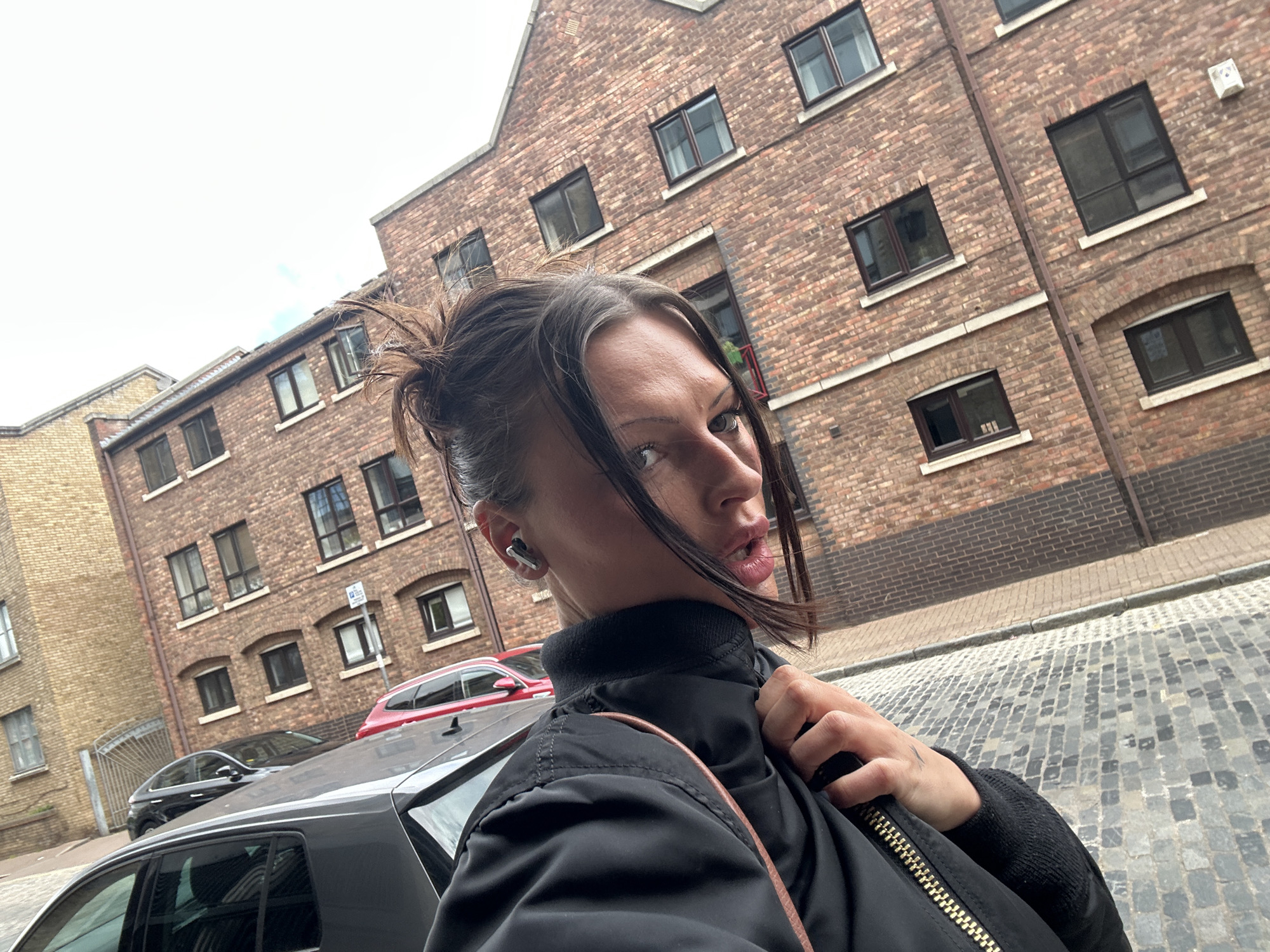 A woman is taking a selfie on a cobblestone street in what appears to be an urban area of London, United Kingdom. She has brown hair pulled back in a messy bun, a fair complexion, and is wearing a black bomber jacket. She has what appear to be earbuds in her ear. In the background are several brick buildings with multiple windows. A red car and a black car are partially visible behind her. The sky is overcast. The street and buildings are weathered, suggesting an older part of the city.