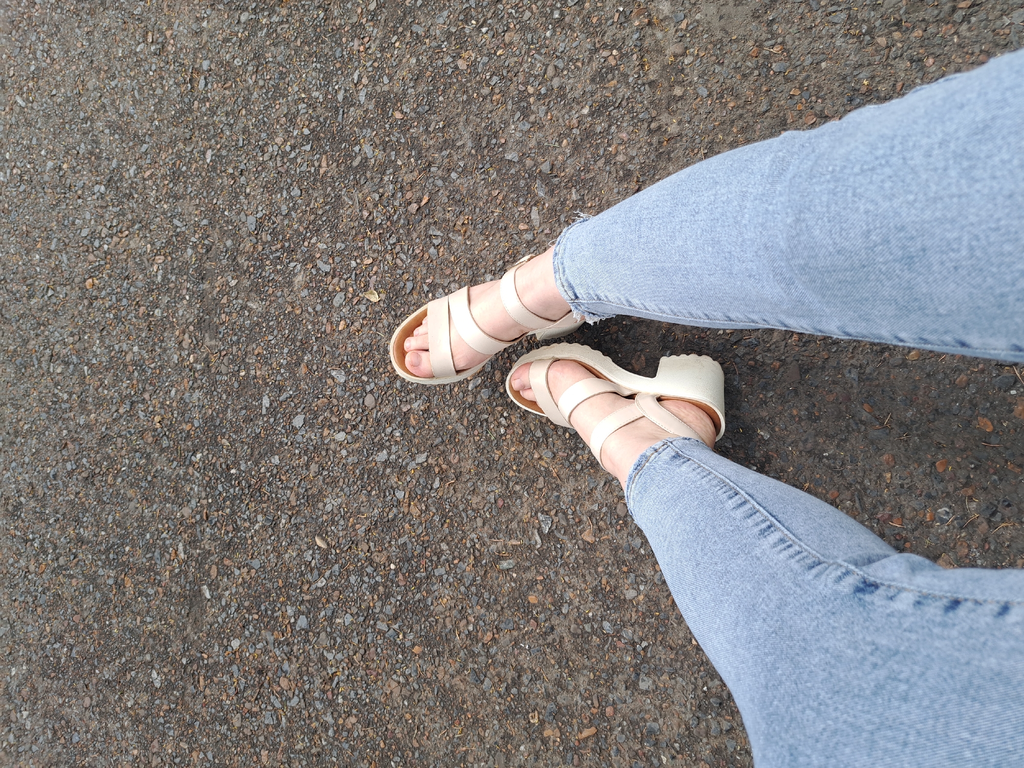This is a top-down, medium shot of a person's legs and feet, wearing light blue jeans and beige heeled sandals, standing on a gravelly asphalt surface. The scene is neutral and appears to be outdoors during the day, possibly in a pedestrian area or sidewalk, given the texture of the ground. The lighting suggests it is not direct sunlight, perhaps overcast or shaded. There is no visible text in the image. The person's stance is relaxed, with one leg slightly in front of the other. The jeans have a slightly frayed hem. The sandals are open-toed with multiple straps and a chunky heel. The overall impression is casual and everyday.