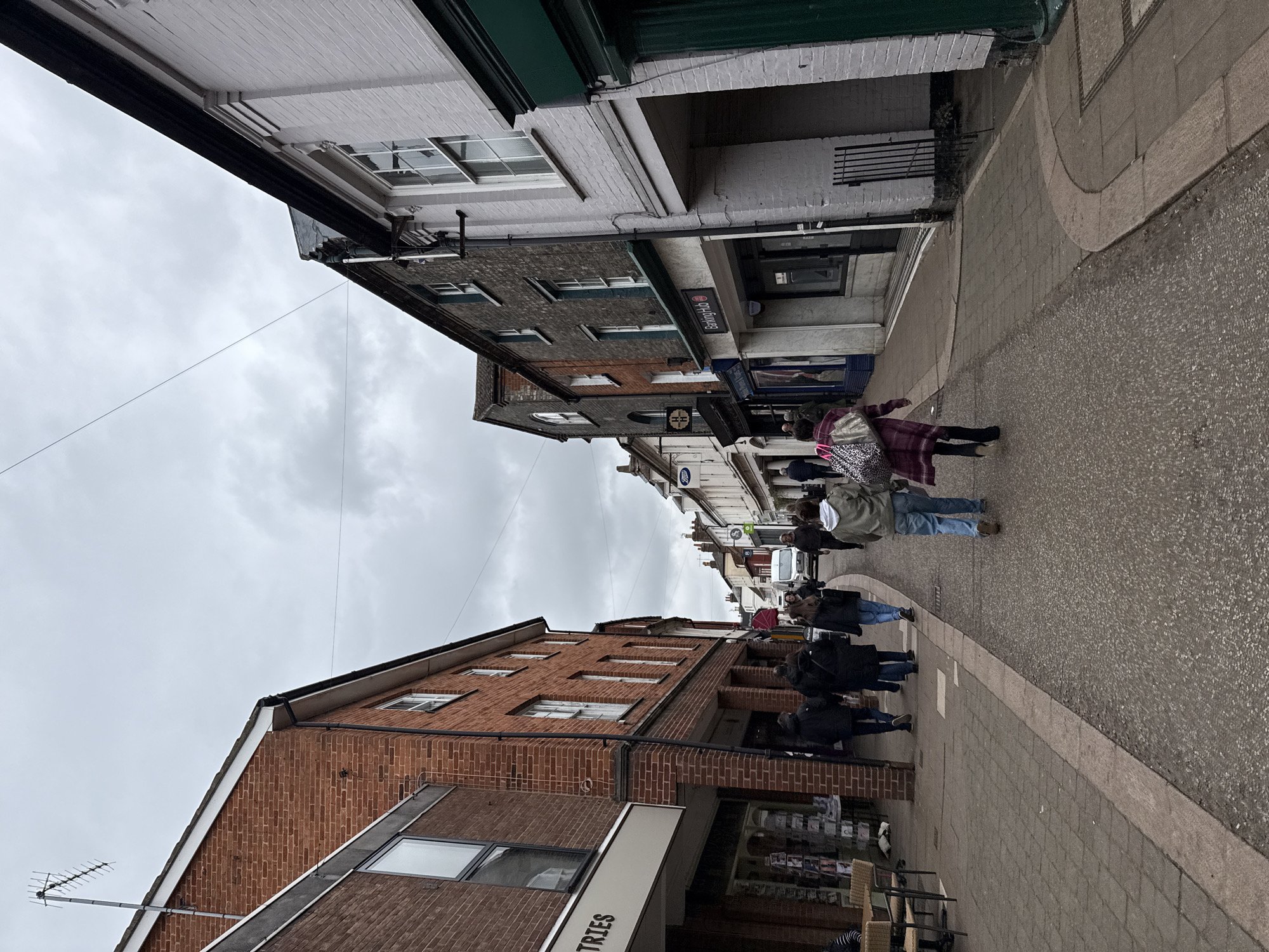 The image depicts a street scene in East Suffolk, United Kingdom. The street is lined with brick and painted buildings of varying heights and architectural styles. Several people are walking along the sidewalk, some in groups. Overhead, a cloudy sky is visible between the buildings. The street surface is a mix of pavement and cobblestone. Storefronts are visible, including one with a sign that partially reads "TRIES."