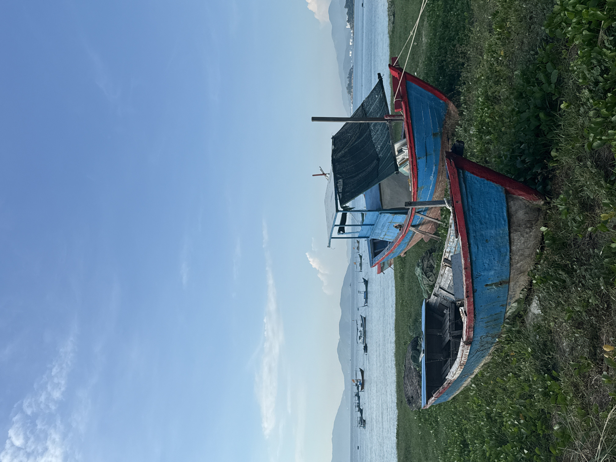 Two weathered, blue and red fishing boats rest on a grassy shoreline facing a calm body of water. The boat in the foreground is older and more dilapidated, with visible wear and tear on its hull. It is angled towards the viewer, with its interior showing wooden planks and debris. The second boat, situated further back and slightly to the right, is larger and appears to be in better condition. It has a blue canopy providing shade over its midsection. Both boats are tied with ropes. In the distance, a line of small fishing boats is visible on the water, with a mountain range forming the backdrop under a clear, pale blue sky with sparse clouds. The scene is likely located in Bac Nha Trang Ward, Vietnam, given the characteristic fishing boats and coastal landscape. There are no people or animals present in the image.