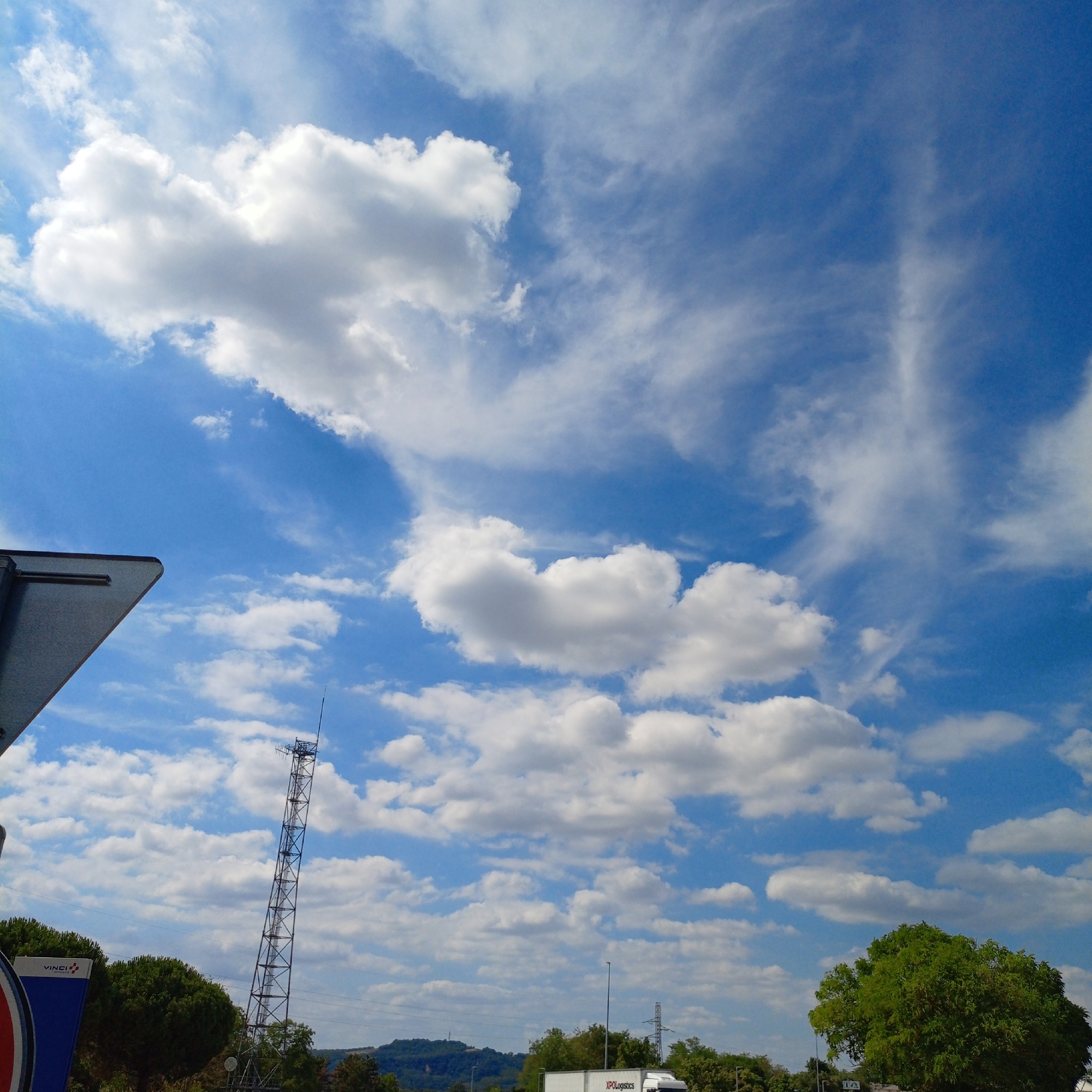 The media file depicts a bright, sunny day with a clear blue sky filled with scattered white clouds. The image captures a wide outdoor scene, likely a roadside or highway area, given the presence of a tall communication tower and a truck with "KPOLogistics" visible on its side. A large green tree dominates the right side of the frame, and another smaller cluster of trees is seen on the left. A triangular signpost is partially visible in the bottom left corner, with a blue panel featuring the text "VINCI AUTOROUTES". The sun appears to be high in the sky, suggesting it is daytime, possibly late morning or early afternoon. The weather appears to be fair and pleasant, with a gentle breeze indicated by the wispy clouds. The overall mood is calm and expansive, dominated by the vastness of the sky.