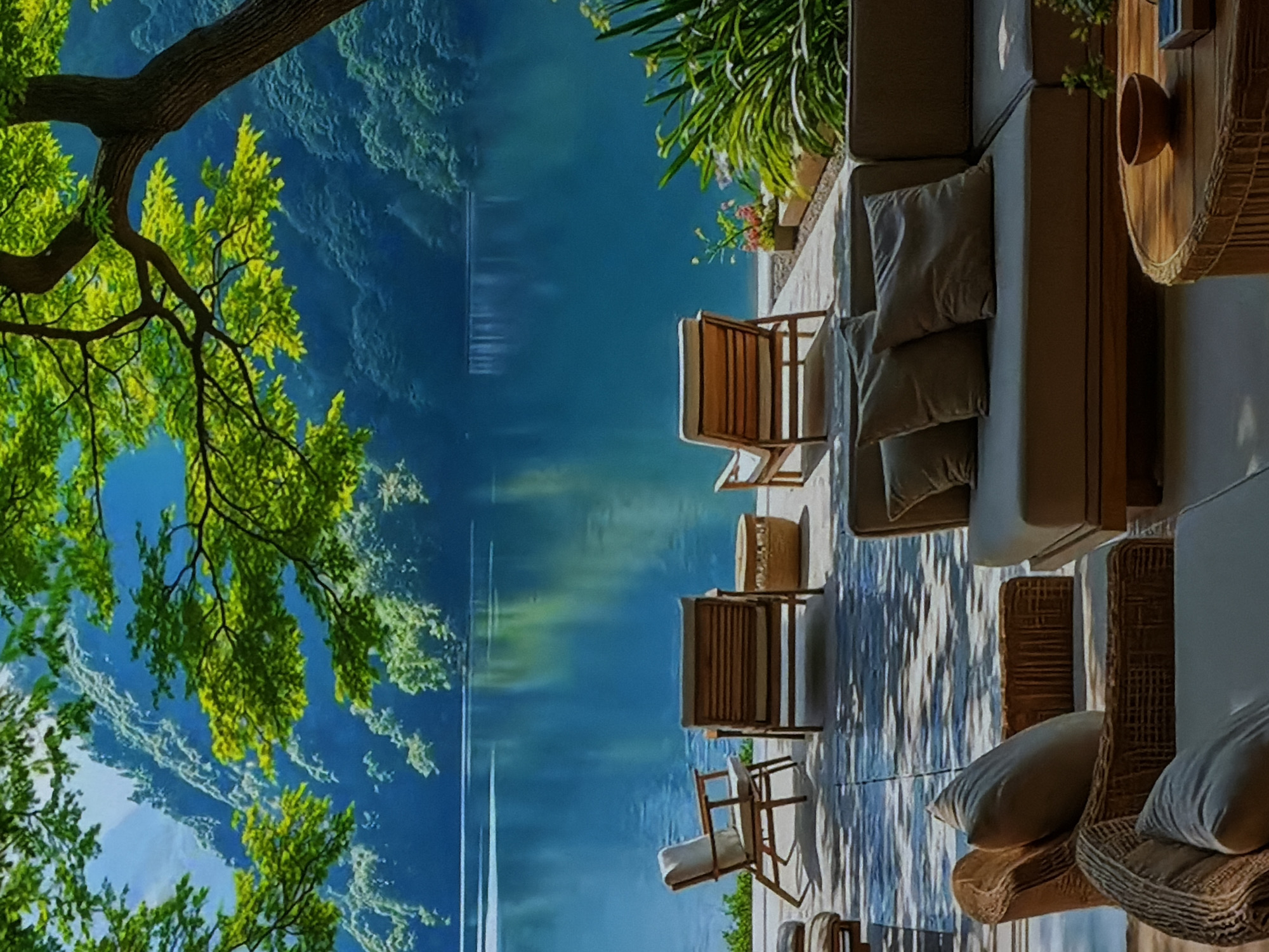 The image depicts a serene outdoor setting, likely a private terrace or patio, offering a panoramic view of a large, calm body of water surrounded by lush, green hills or mountains. The photo is oriented vertically, with the natural landscape dominating the left side and the man-made patio on the right.
In the immediate foreground, on the left, a large tree branch with vibrant green leaves extends into the frame, partially obscuring the distant view and casting dappled light. The thick, dark brown trunk and branches of the tree are prominent.
On the right side of the image, the patio area is furnished with comfortable outdoor seating. Several sections of what appear to be large, cushioned sofas or lounge chairs with light-colored cushions and pillows are arranged on a light-toned paved surface. Two wooden-framed chairs with light cushions are also visible, facing the water. A small, round, woven side table with a light brown bowl or pot rests near one of the sofas. Another small, woven stool or table is seen between the wooden chairs. Green foliage and small flowering plants in planters add to the natural aesthetic of the patio.
The background features a vast expanse of still, blue-green water that perfectly reflects the distant green landscape and the sky, creating a sense of depth and tranquility. The reflections on the water show faint outlines of what appear to be distant structures or landforms, as well as the sky.
The overall scene suggests a peaceful daytime setting, with clear weather and abundant natural light. There are no people visible, emphasizing the quiet and undisturbed nature of the environment. The atmosphere is one of relaxation and connection with nature. There is no visible text in the image. Given the location context, this tranquil scene is situated in Sofiivska Borshchahivka, Ukraine.