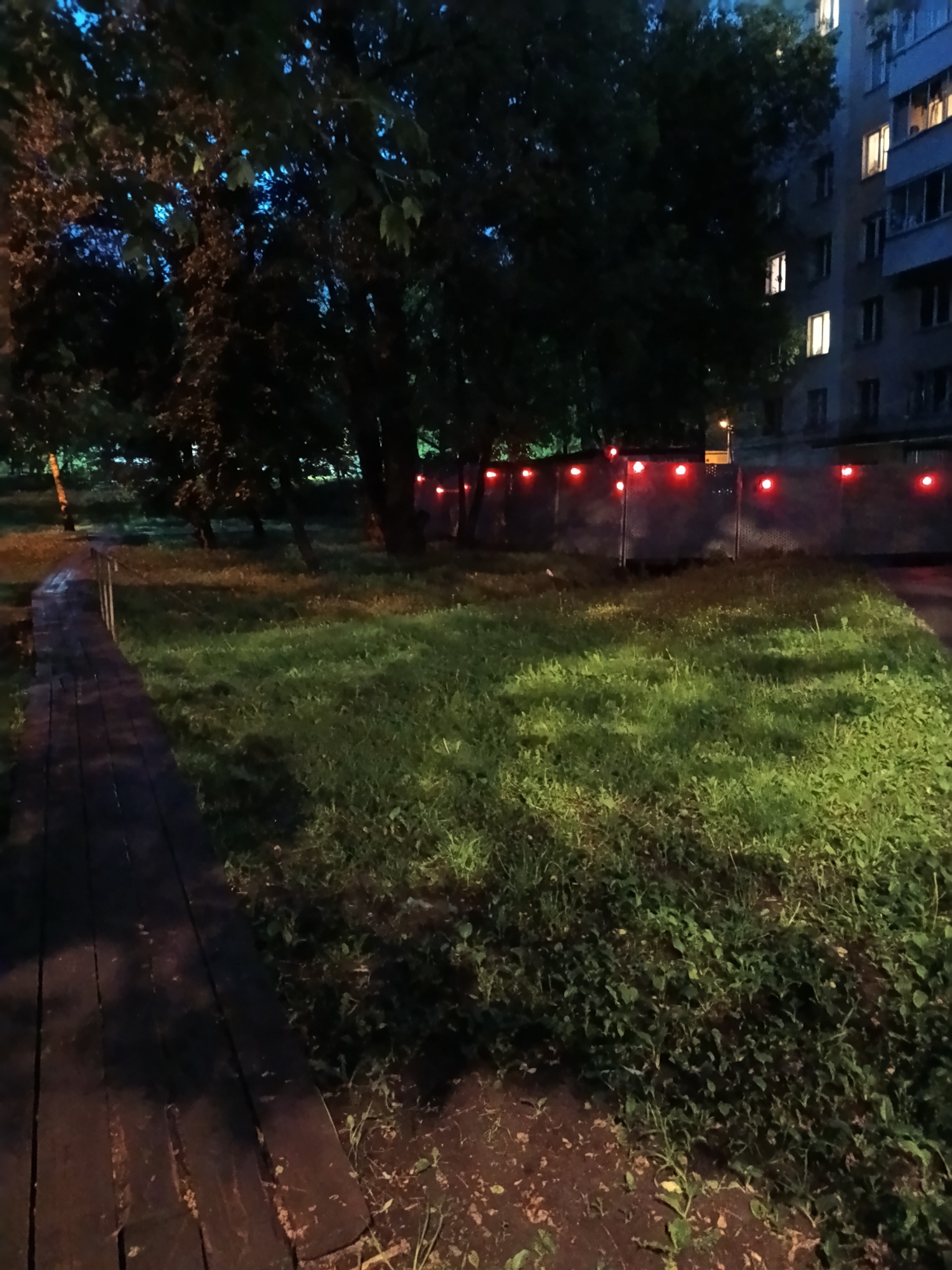 The image depicts a dimly lit park at night in Moscow, Russia. A wooden boardwalk stretches from the lower left corner into the center of the frame, bordered by grass and foliage. A line of trees stands in the midground, partially obscuring a tall fence with evenly spaced red lights. A residential building is visible in the background, with several illuminated windows. The lighting suggests it's either dusk or a poorly lit area after dark. The overall mood is quiet and somewhat eerie due to the low light and red lights along the fence. There are no people visible in the frame.