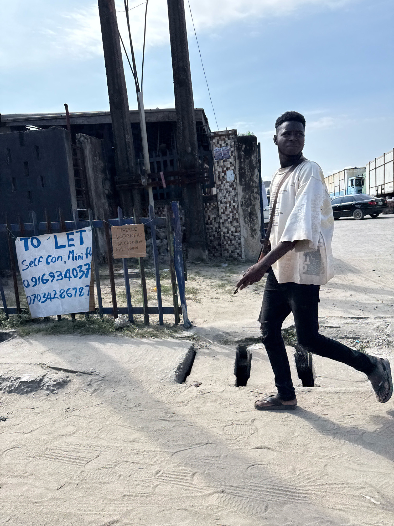 A young Black male wearing a loose-fitting white t-shirt, ripped black jeans, and brown sandals is walking to the right across a dusty, unpaved street. He is looking over his left shoulder. In the background, there are several utility poles, a dark-colored building with a corrugated metal roof, and parked trucks. Two signs are visible on a blue metal fence in the foreground. The sign on the left, written in blue ink on white paper, reads "TO LET Self con, Mini Fl 0916934037 07034286787". The sign on the right, written in black ink on a brown cardboard, reads "MALE WORKERS NEEDED HERE APPLY WITHIN". This scene is in Ado, Nigeria.