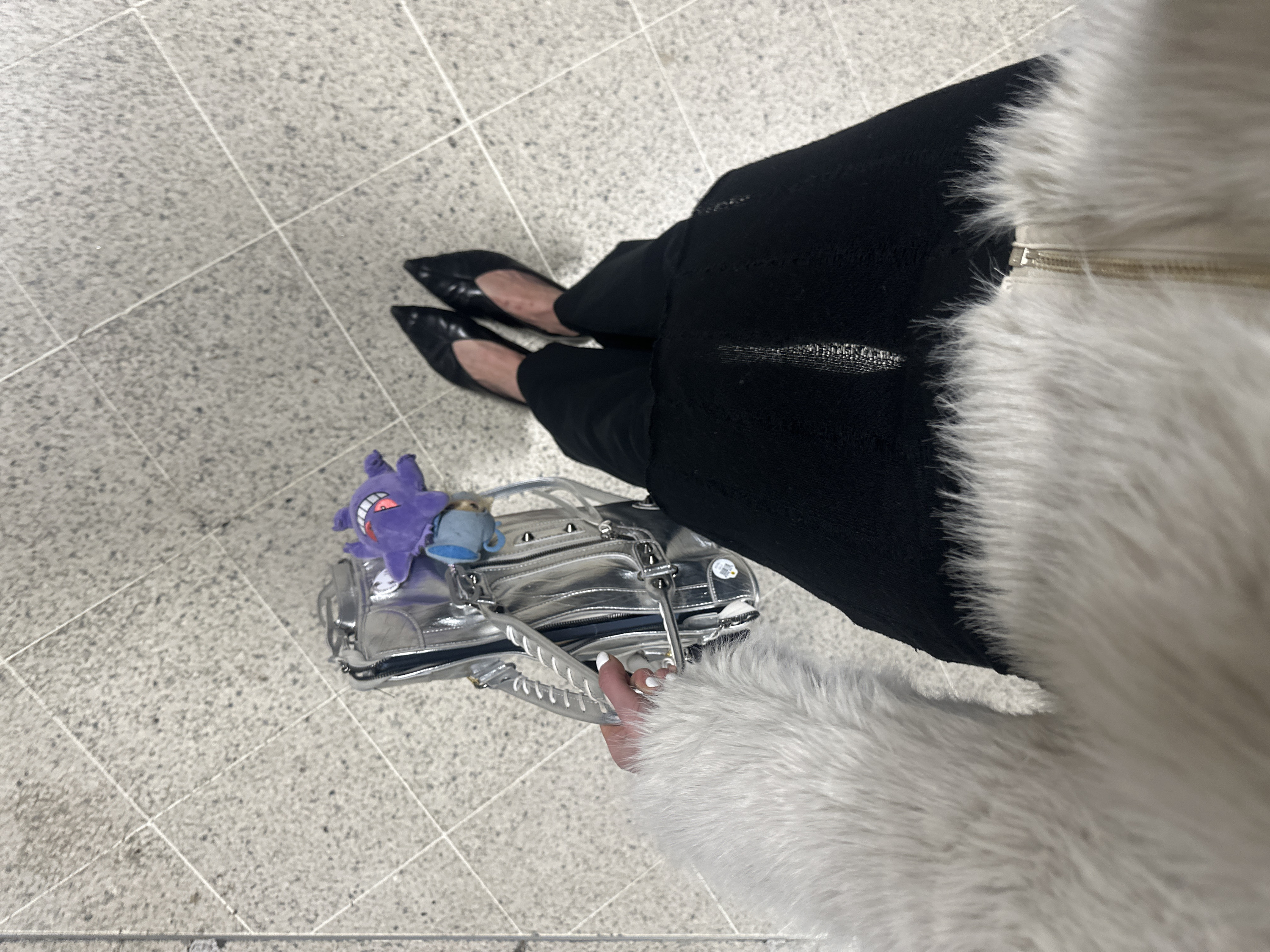 A first-person, high-angle view captures the lower torso and legs of an individual standing on a light grey, speckled tiled floor with white grout lines, indicative of an indoor public space. The individual wears a white faux fur coat or jacket with a gold zipper, black trousers or a skirt, and black pointed-toe flat shoes. Their left hand, featuring white nail polish, holds a metallic silver handbag or backpack with braided white details on the strap. Attached to the bag are two plush keychains: a purple Gengar character with an open mouth and red interior, and a smaller light blue object resembling a teacup. A sticker partially visible on the silver bag displays "LHR," identifying the location as London Heathrow Airport in London, United Kingdom.