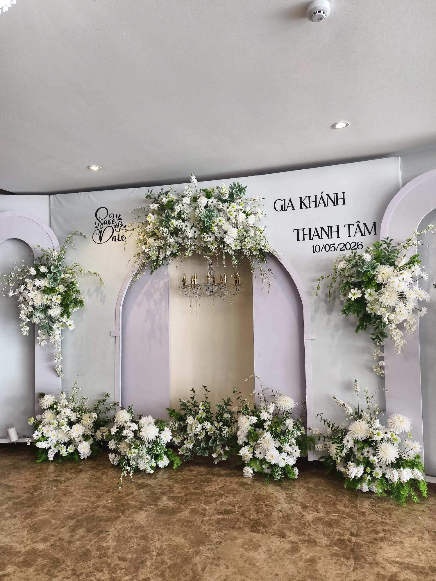 A wedding backdrop is set up in Bac Nha Trang Ward, Vietnam. The backdrop is a light gray wall with two stylized arches on either side of a larger, central arch. The wall has the phrases "Save the Date", "Gia Khanh", and "Thanh Tam 10/05/2026" printed on it in black lettering. The arches are decorated with arrangements of white flowers and greenery. A small, ornate chandelier hangs within the central arch. The floor is a polished brown marble.
