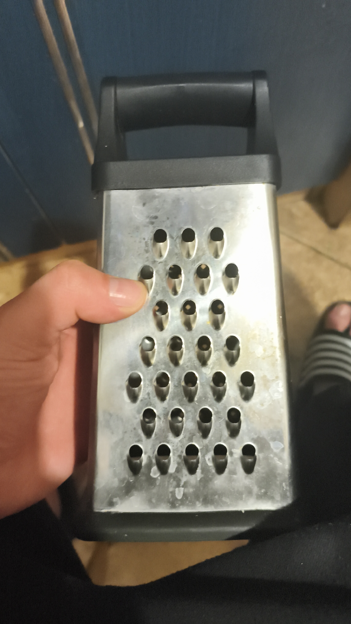 A close-up photograph depicts a rectangular, box-shaped grater made of silver metal with a black plastic handle and base. The grater has multiple small, round grating holes arranged in a triangular pattern. A human hand is partially visible on the left, with a finger touching the grater's surface. The grater appears to be in use, with some residue visible in and around the holes.
The background is blurry and suggests an indoor setting, with a blue cabinet partially visible on the left, a wooden floor on the top right, and clothing at the bottom. A foot wearing a striped slider is also visible.
The image does not provide information to ascertain if the photo was taken in Nowy Tomyśl, Poland.