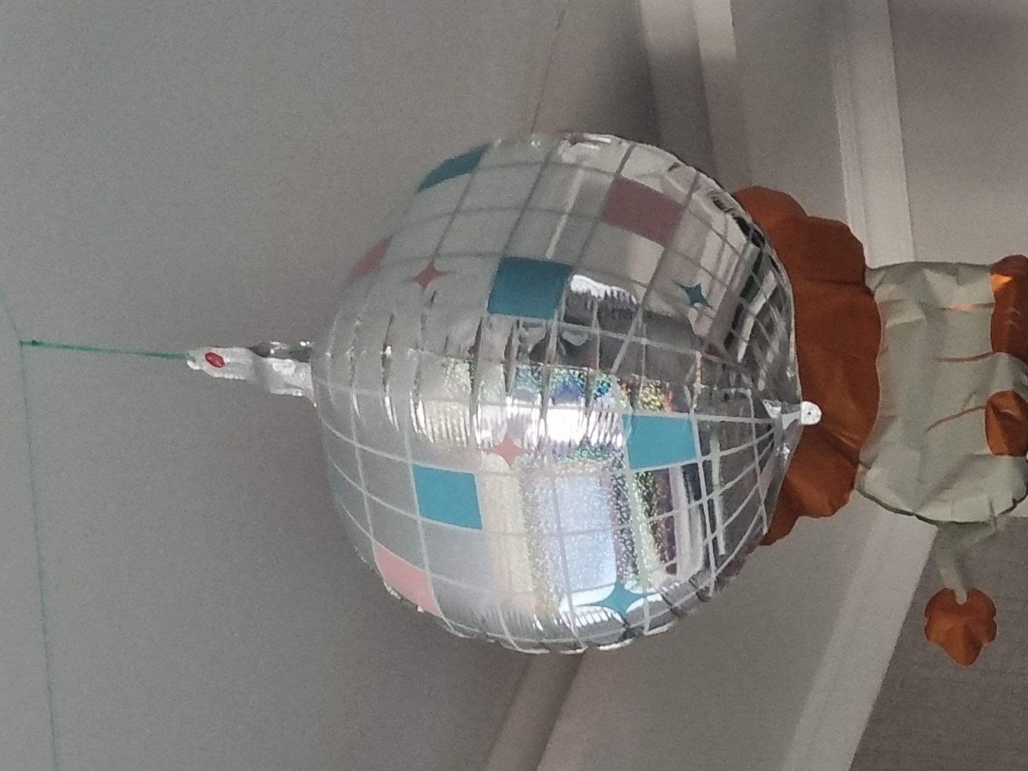 The image shows a silver disco ball with pink and blue square accents and star shapes, suspended from a green string attached to a ceiling. Below the disco ball, the top portion of an inflatable figure is visible, appearing to have orange arms and a white body. The background is a grey ceiling with some textured molding. The scene is indoors. No information is provided about the city of Samara, Russia.