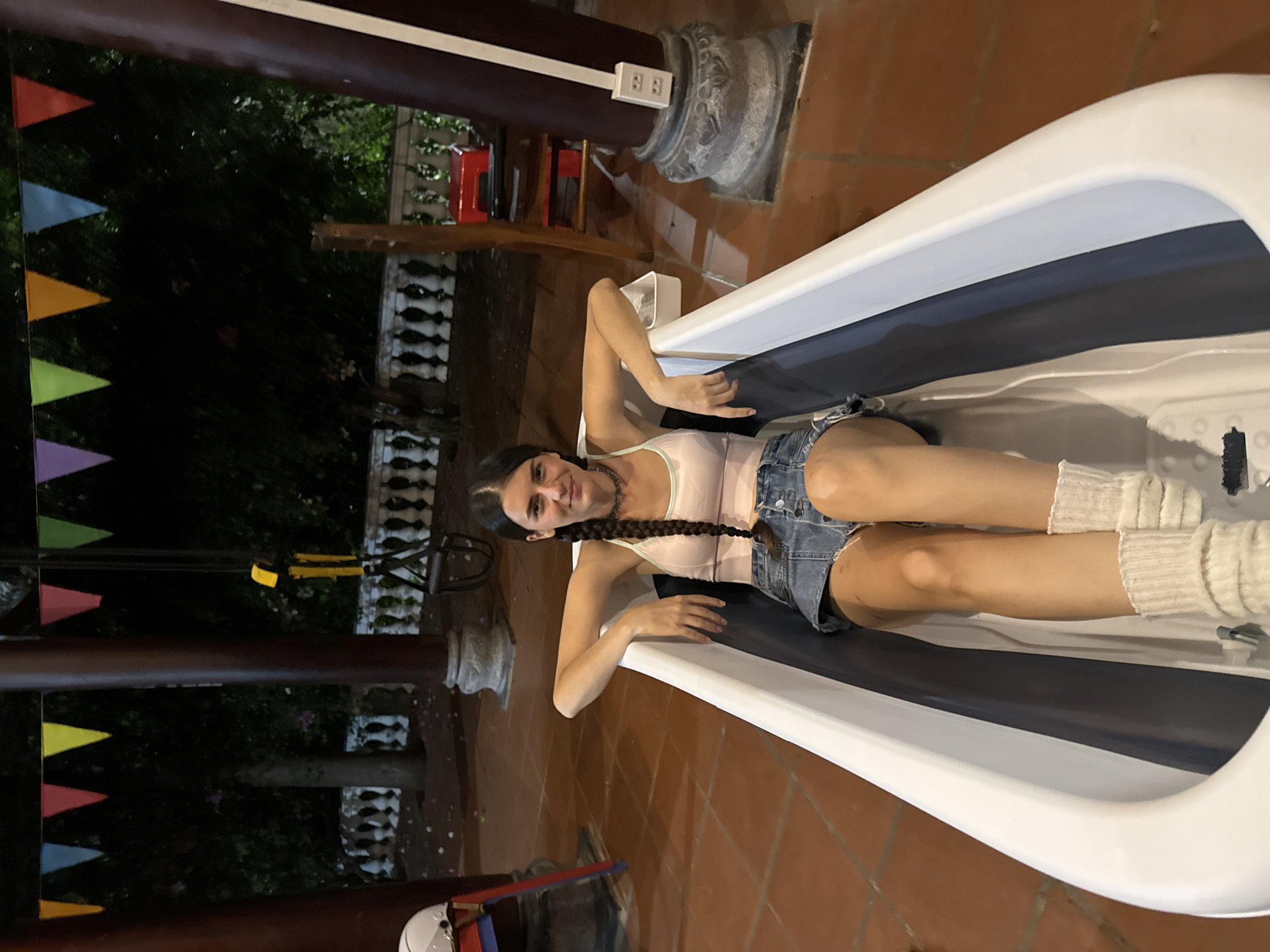 A medium shot captures a woman lying in a white, blue, and black bathtub outdoors. She is wearing a light pink top, denim shorts, and white socks. She has dark hair braided down her back and is smiling.
The bathtub is positioned on a tiled patio. In the background, there is greenery and a structure with decorative columns. String lights with colorful triangular flags are hung overhead. A helmet is also visible near the bathtub.
The location is likely Mũi Né, Vietnam, based on the image’s metadata.