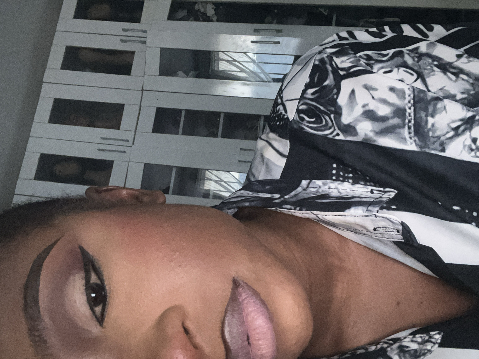 A close-up, partial portrait shows a Black woman's face and upper torso. She is wearing a black and white patterned shirt. Her makeup includes winged eyeliner, eyeshadow, and blush. Her lips are painted a light pinkish-purple color. In the background, white cabinets with glass doors are stacked. The lighting is dim. The image does not provide any information about the location being the city of Abuja, Nigeria.