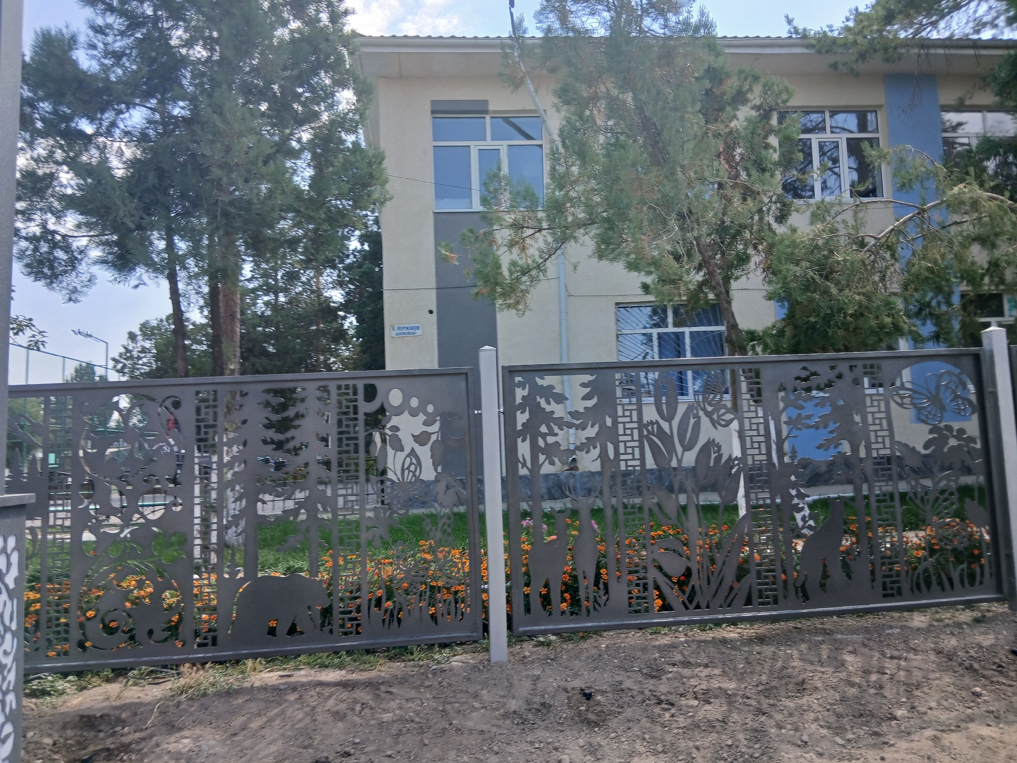 The image shows a metal fence with cutouts of trees, animals, and flowers. The fence is in front of a building that appears to be a school or educational institution. The building has a beige facade with blue accents and several windows. Green trees with sparse foliage are visible behind the fence and around the building. Orange flowers are planted in front of the fence. The sky is visible in the background and is partly cloudy. The overall scene suggests an outdoor setting in a city, possibly a park or school grounds. The metal fence is the primary focus, with its intricate design. The presence of a street sign on the building, although partially obscured, hints at a specific urban location. It is located on a street in Talas, Kyrgyzstan.